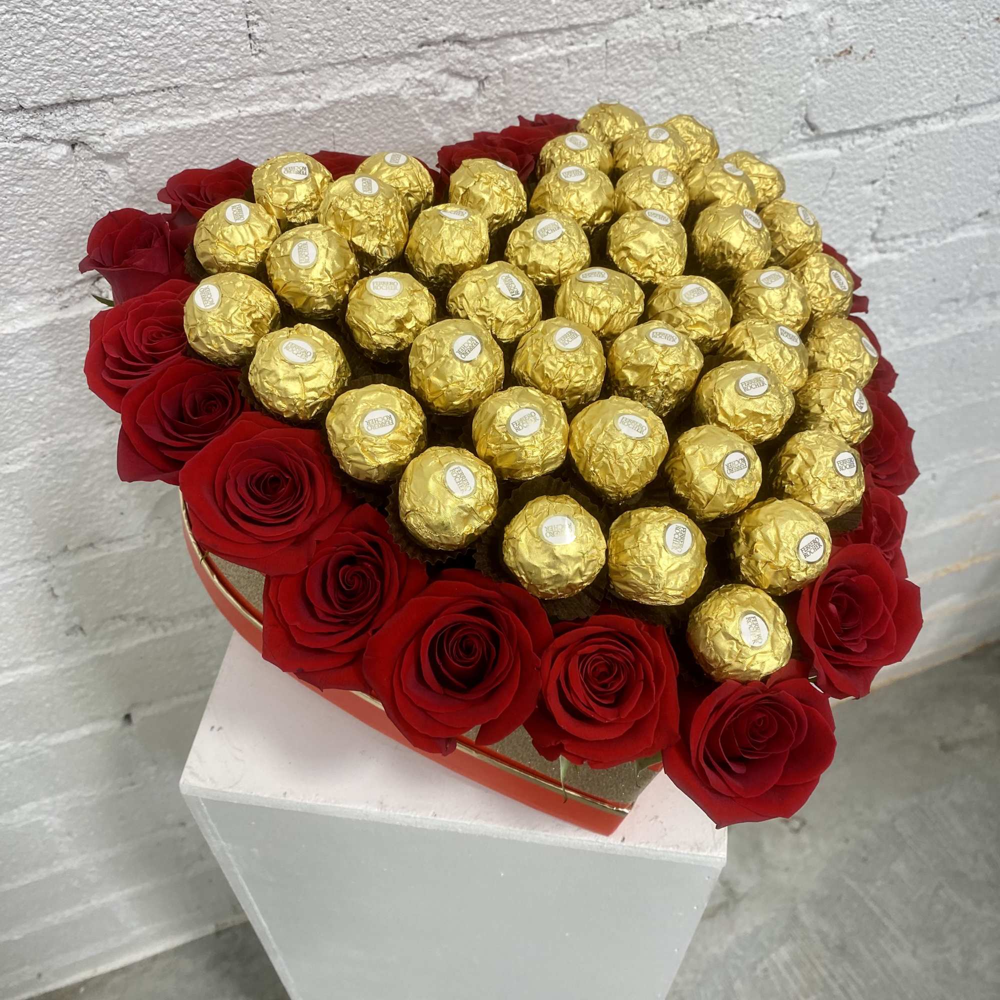 Red roses arranged around gold-wrapped chocolates in a heart-shaped box.