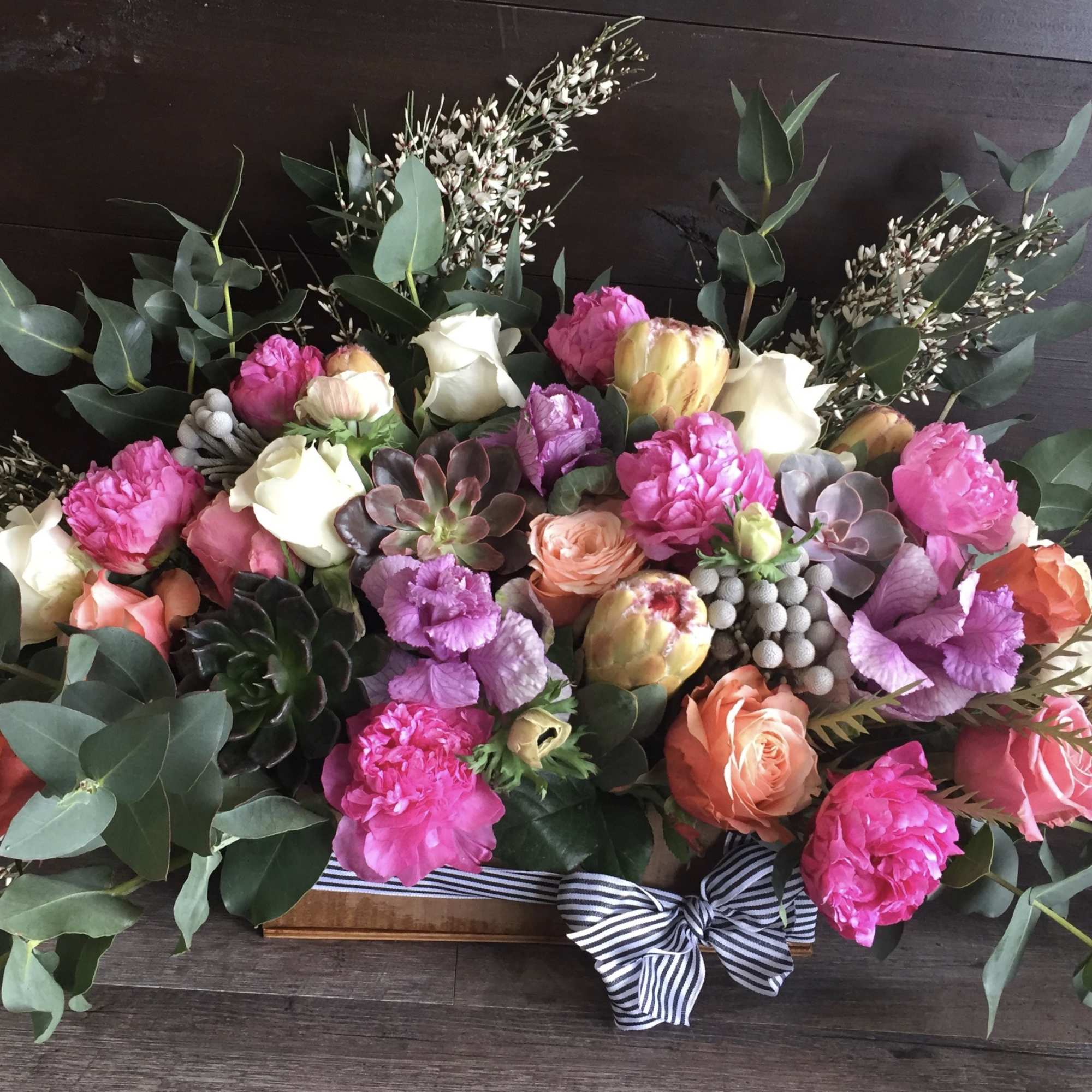 Low rectangular arrangement of pink peonies, mixed roses, succulents and protea in a wooden box with a striped bow