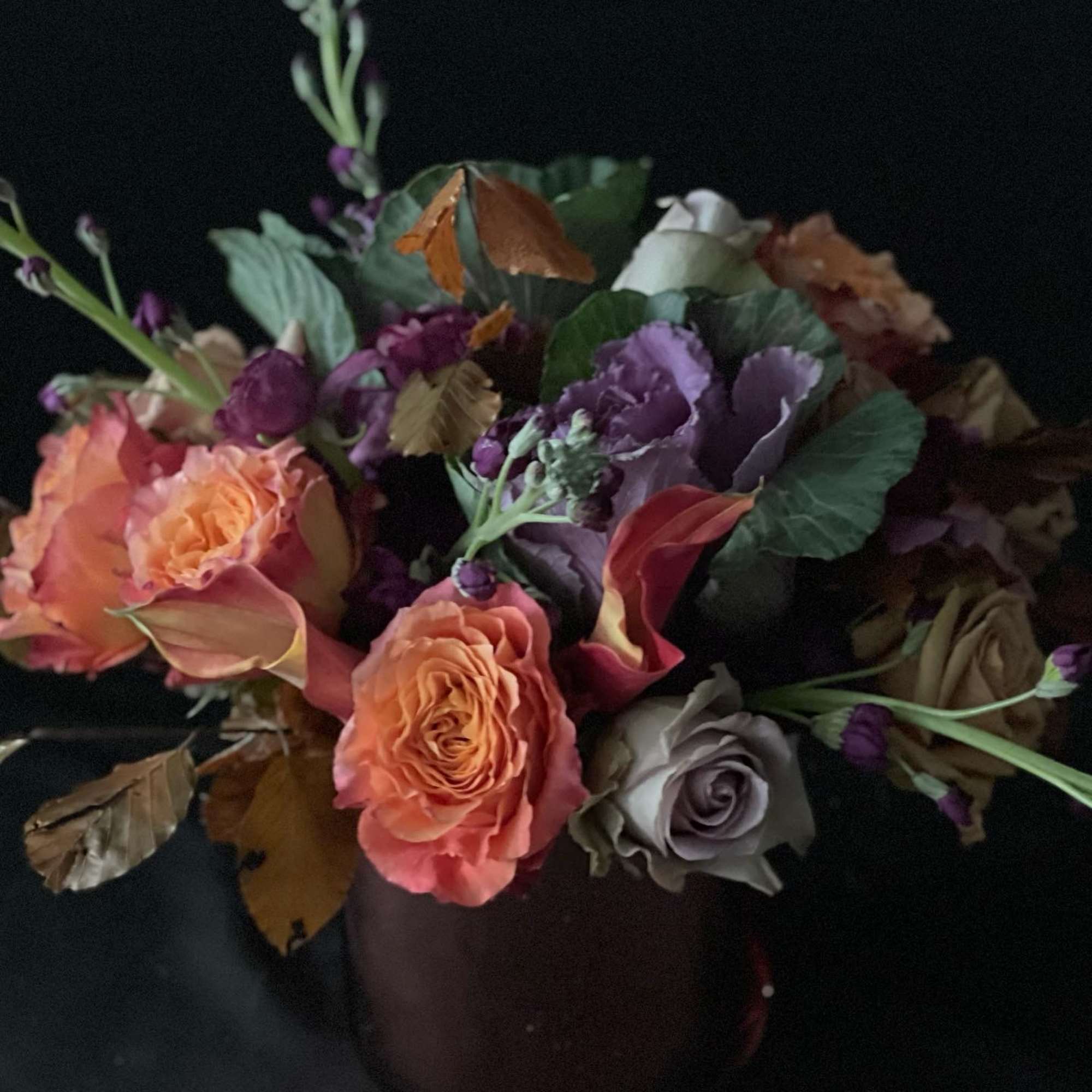 Low arrangement of orange and lavender roses with purple blooms in a dark vase against a black background