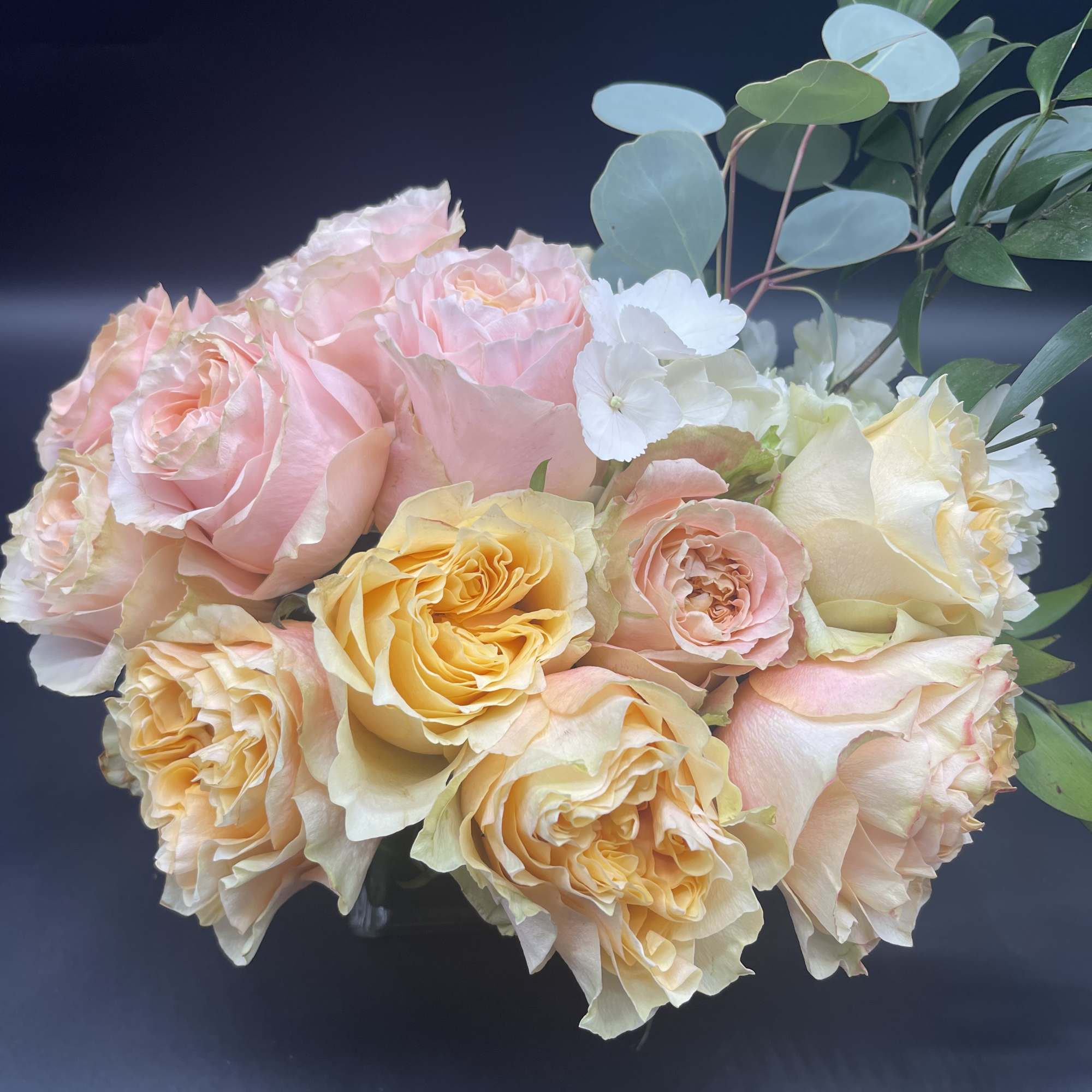 Low arrangement of peach and pink roses with white blooms in a clear glass vase against a dark background