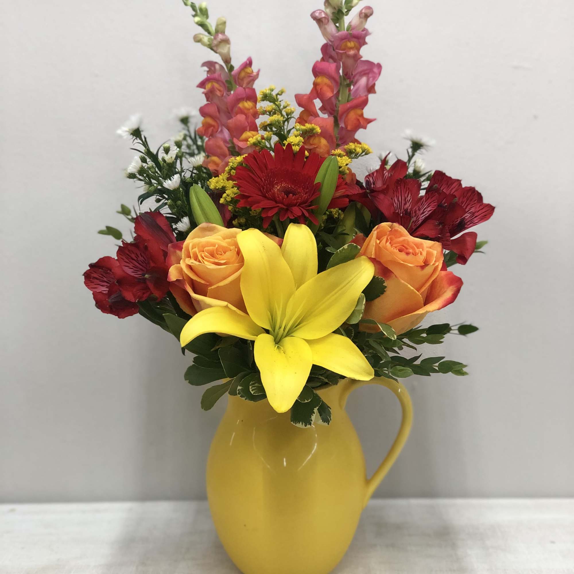 Colorful bouquet in a yellow pitcher vase with roses, lilies, and snapdragons