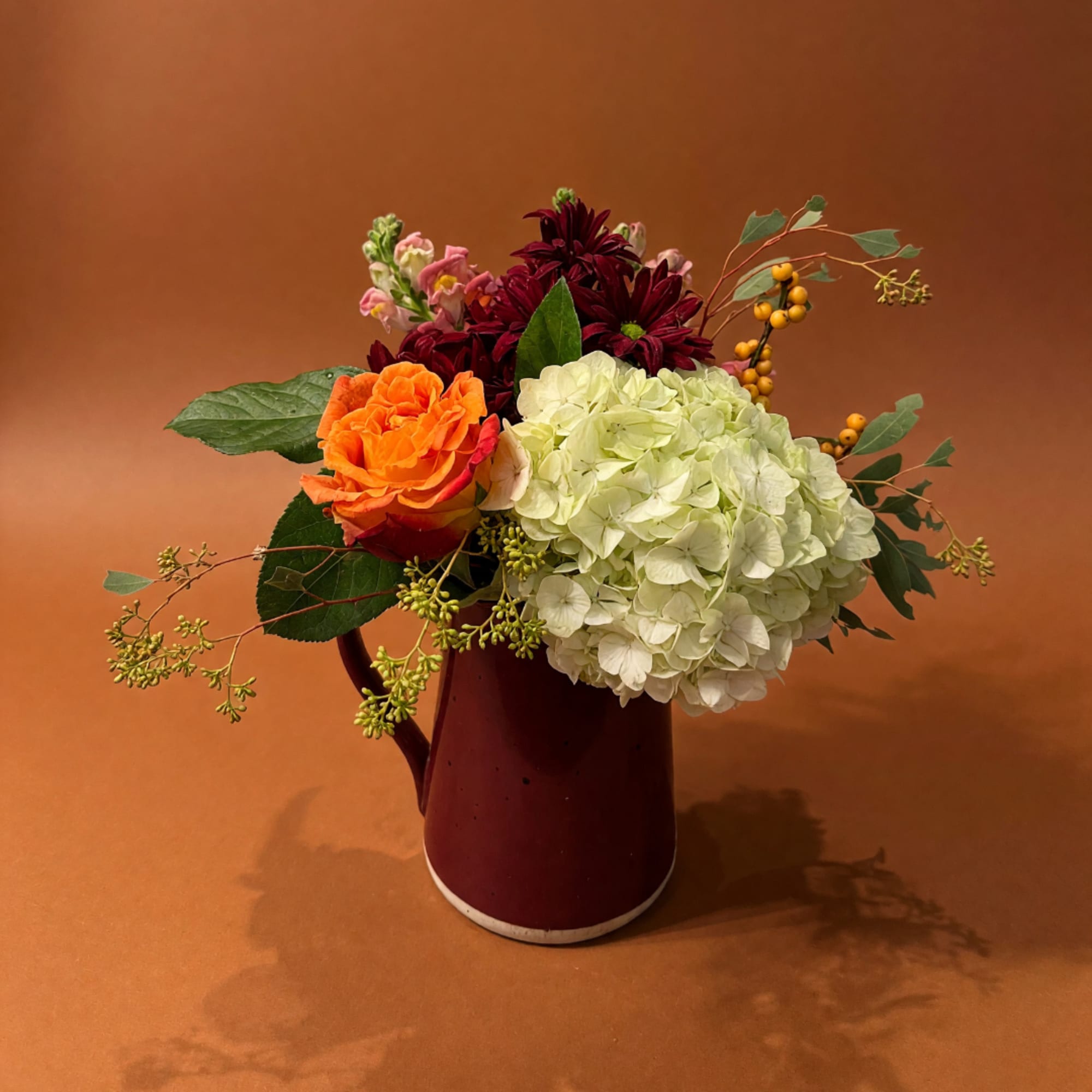 Mixed bouquet with orange rose, white hydrangea, and burgundy blooms in a red pitcher