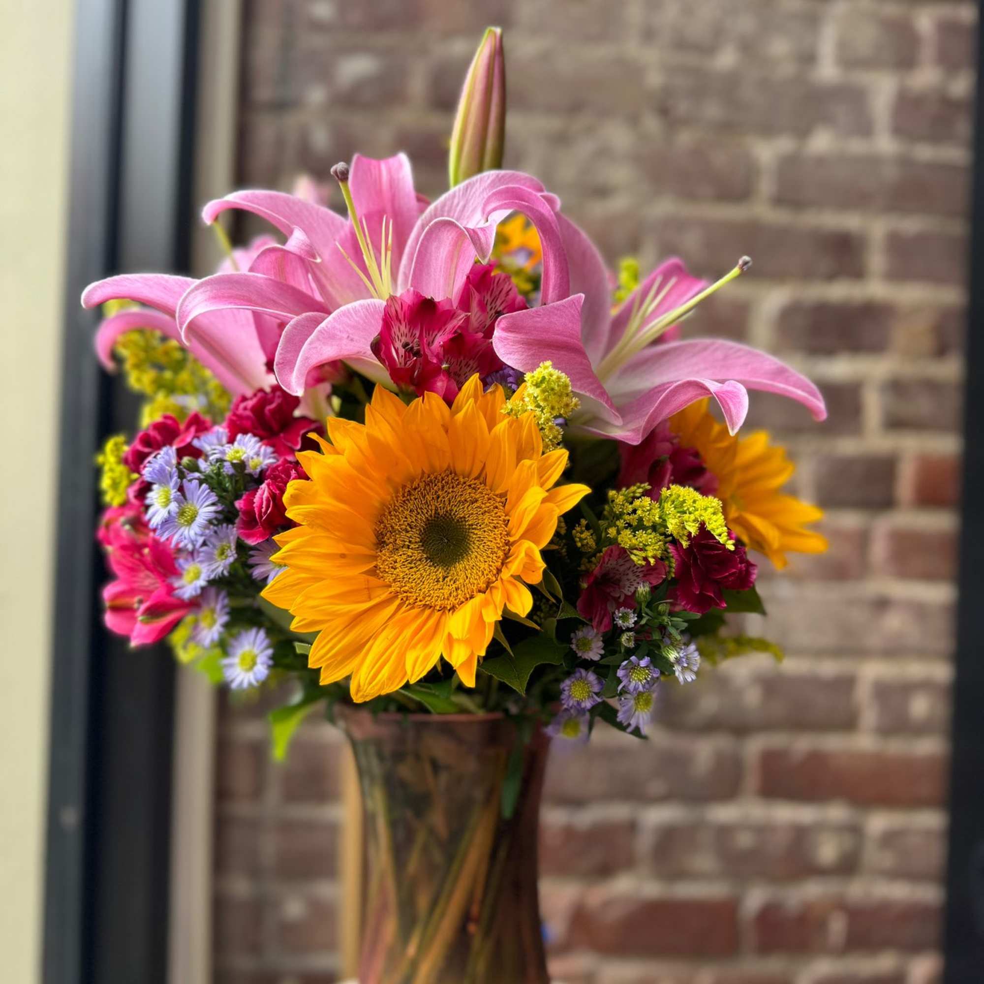 Tall bouquet of yellow sunflowers and pink lilies in a bronze-toned glass vase