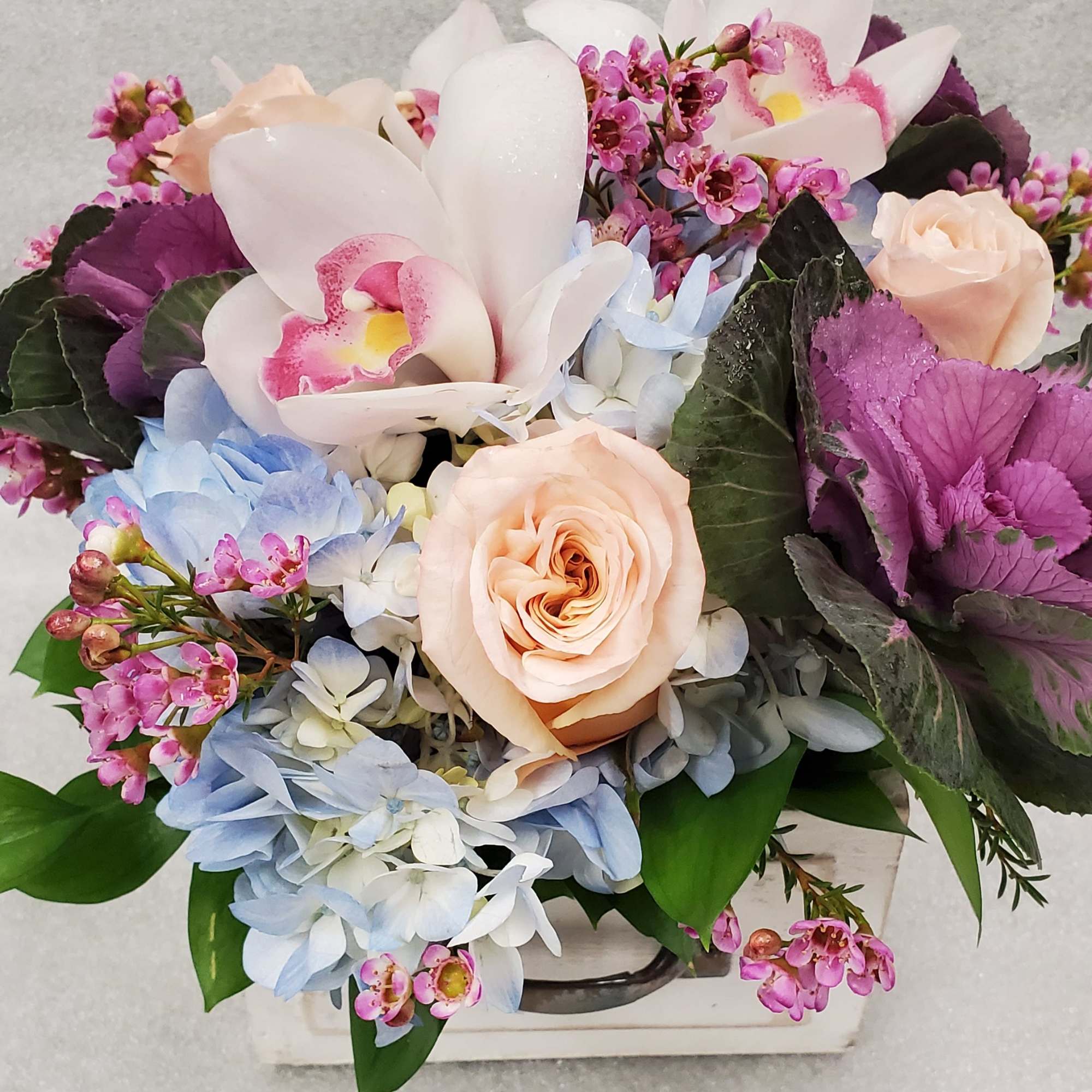 Mixed bouquet with pale roses, orchids, and blue hydrangeas in a white container