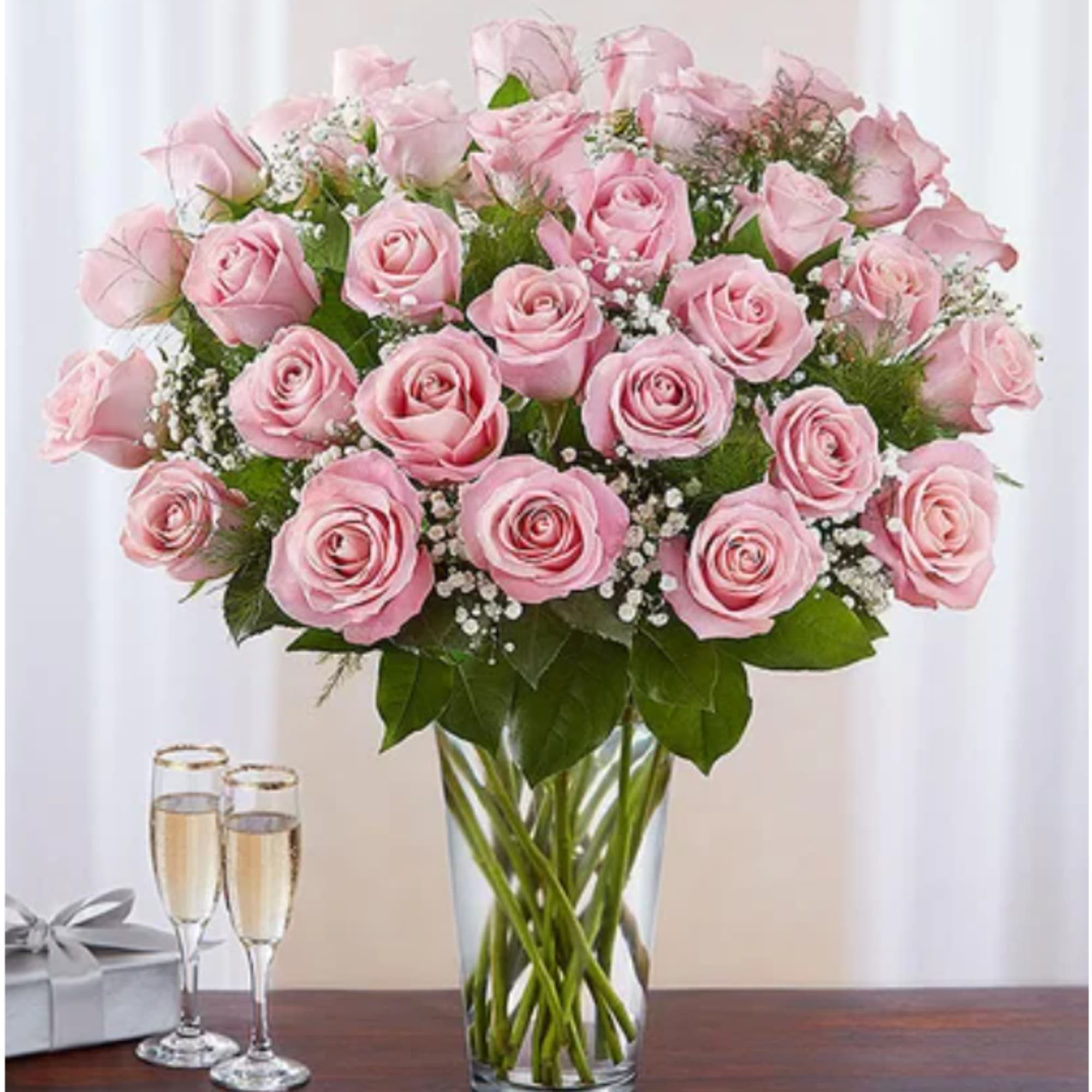Pink roses arranged in a clear glass vase beside two champagne flutes and a gift box.