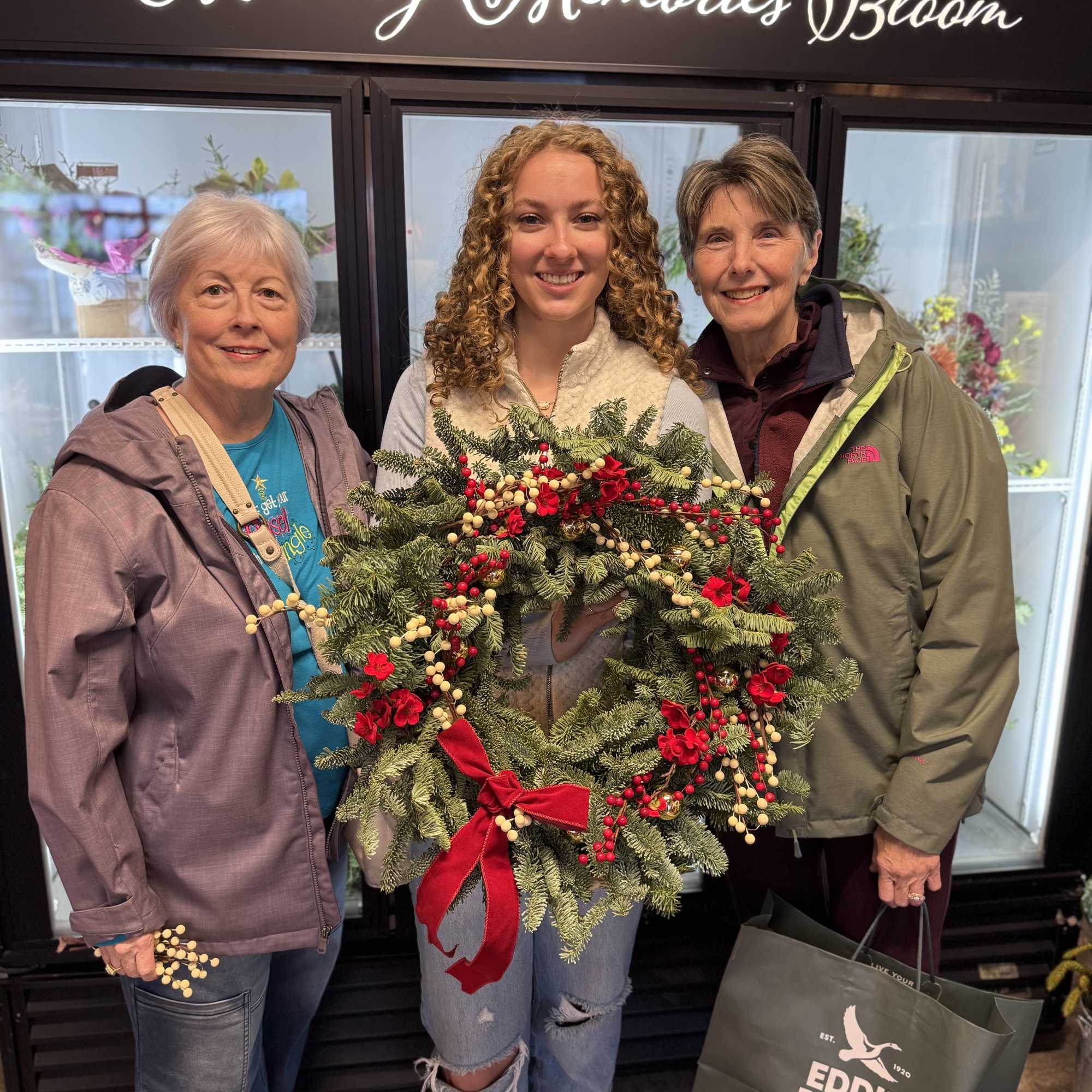Three women hold a decorated evergreen wreath with red berries and a red bow.