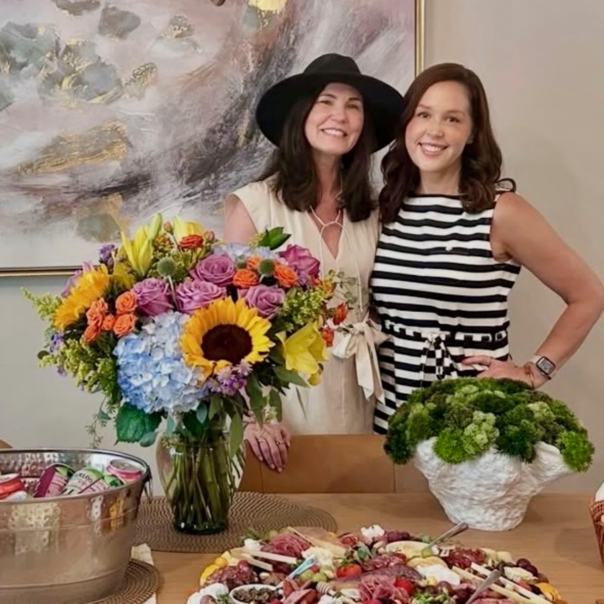 Two women stand behind a colorful bouquet and a green potted arrangement on a table.