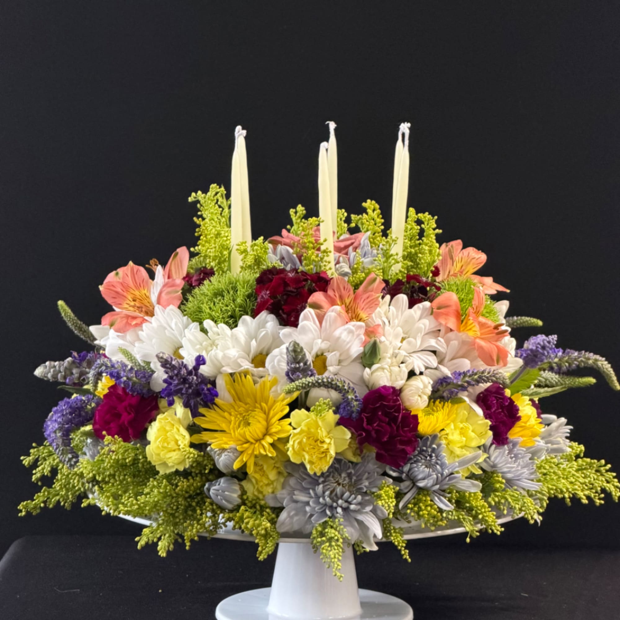 Colorful floral centerpiece with four tall white candles in a white pedestal bowl