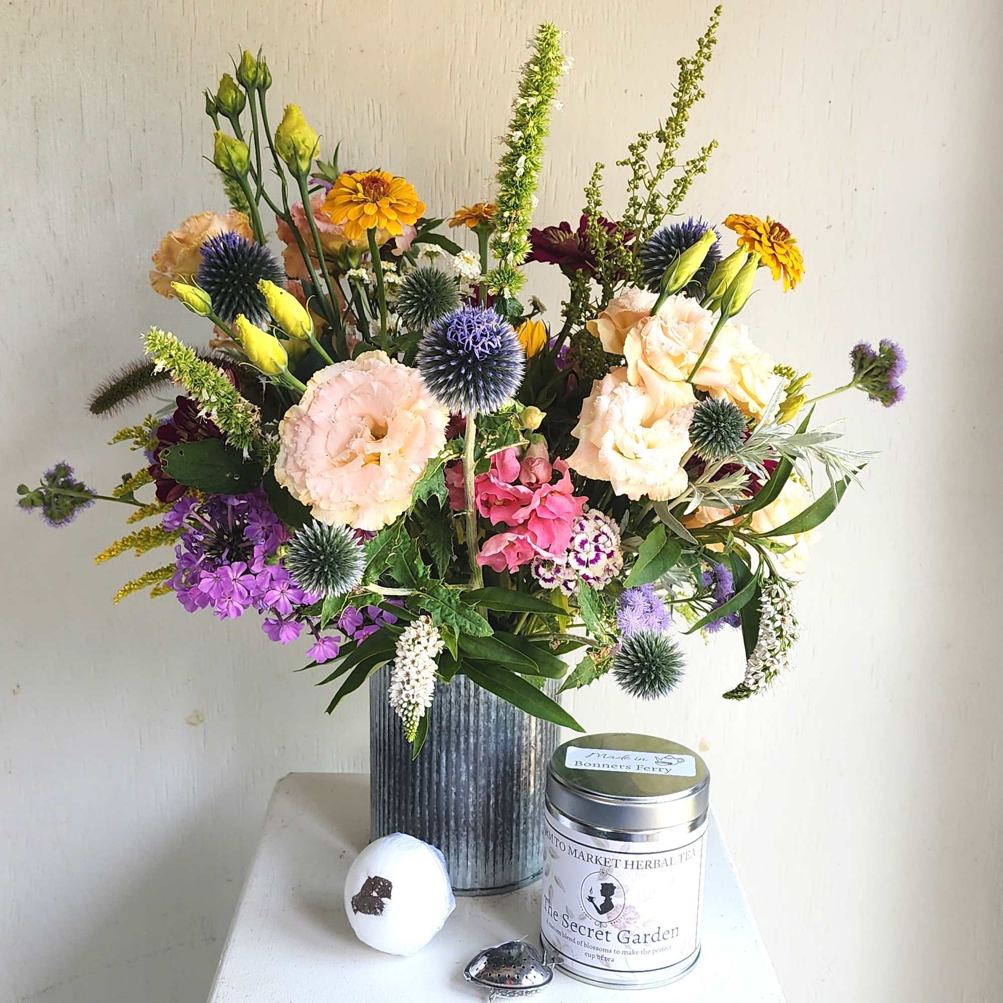 Mixed bouquet in a ribbed metal vase with a tea tin and bath bomb beside it.