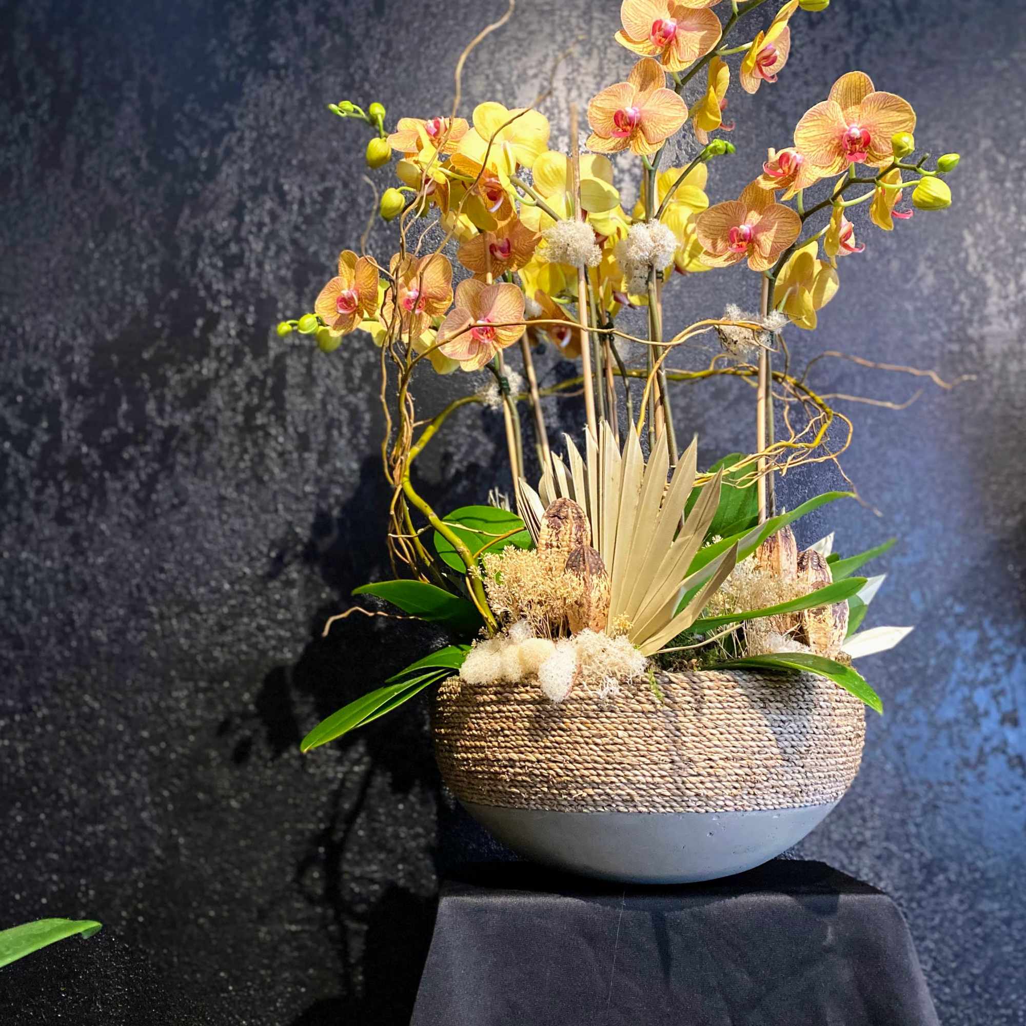 Tall yellow phalaenopsis orchid arrangement in a low woven bowl with dried accents.
