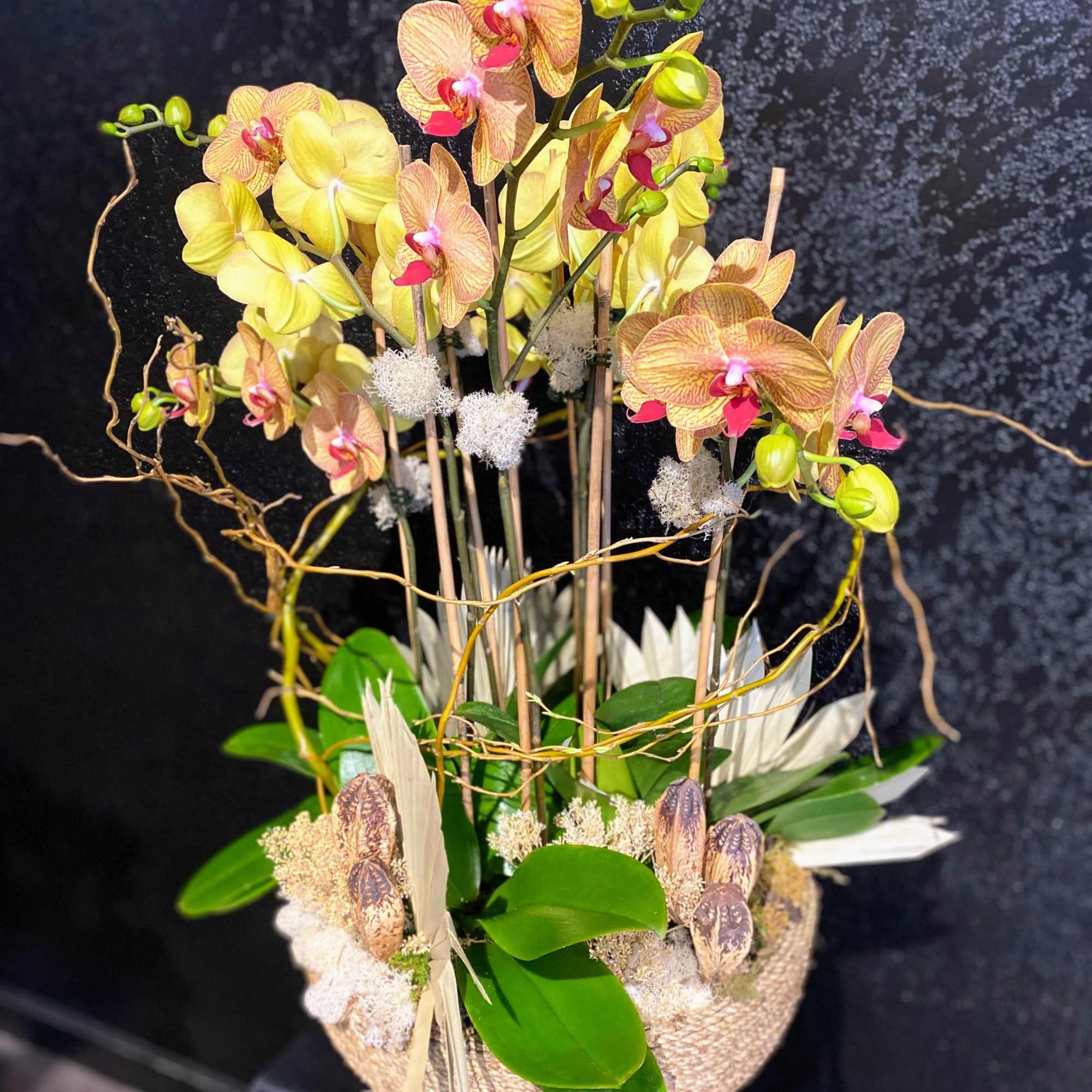 Tall arrangement of yellow phalaenopsis orchids in a low woven basket with green leaves and dried accents