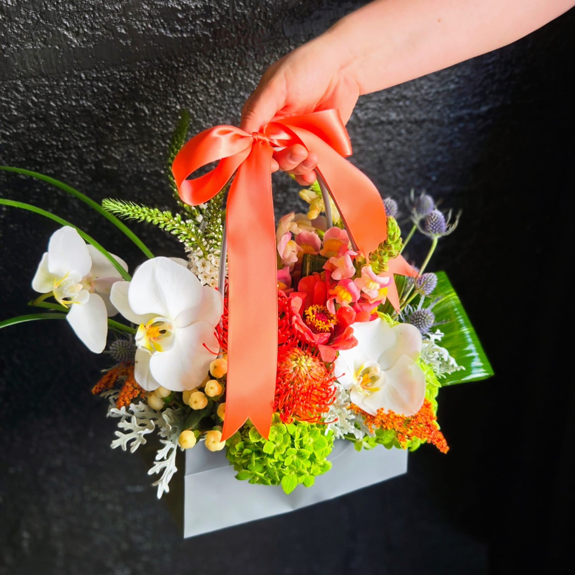 Handheld floral basket with white orchids and bright mixed blooms tied with a coral ribbon