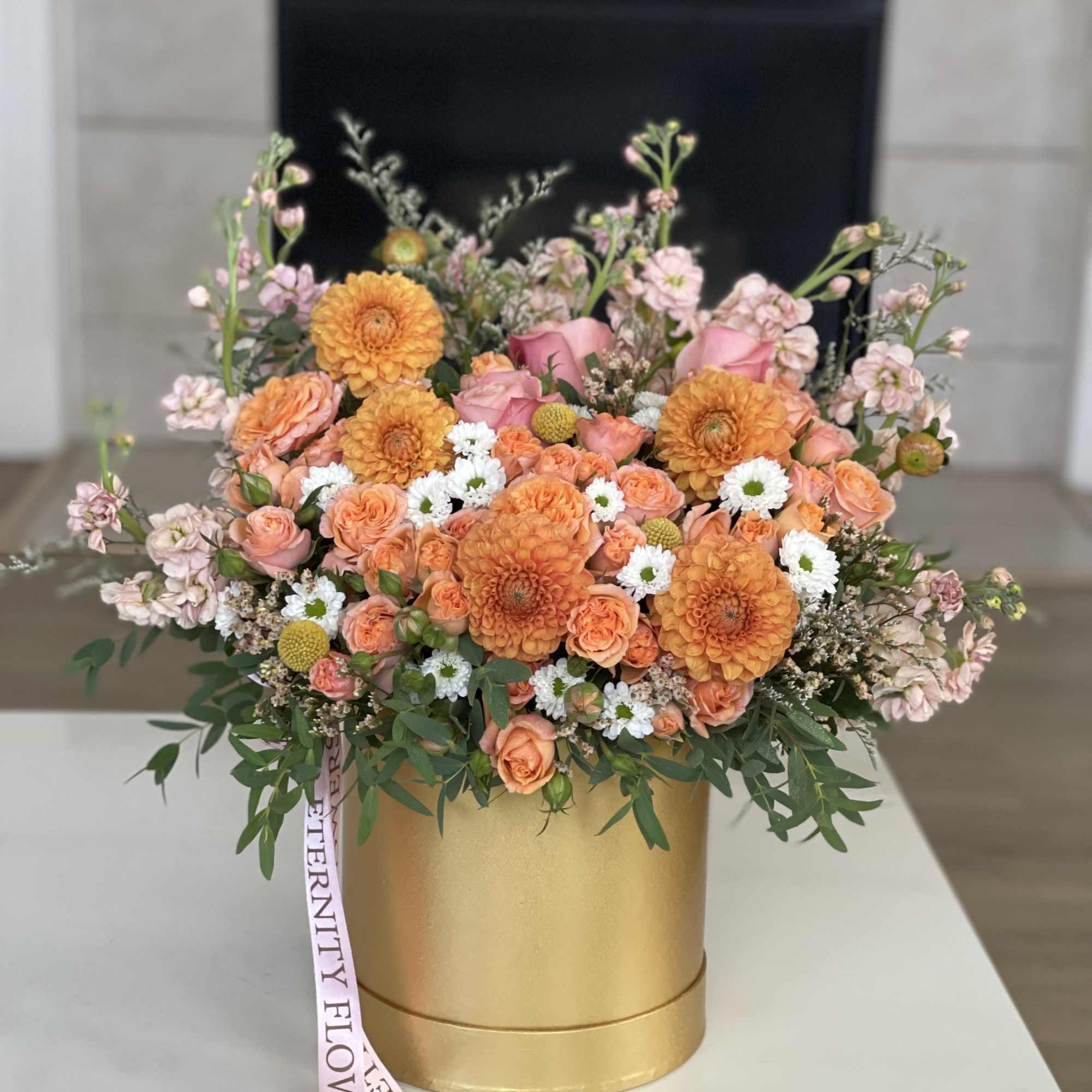Orange and pink floral arrangement in a gold box with a ribbon