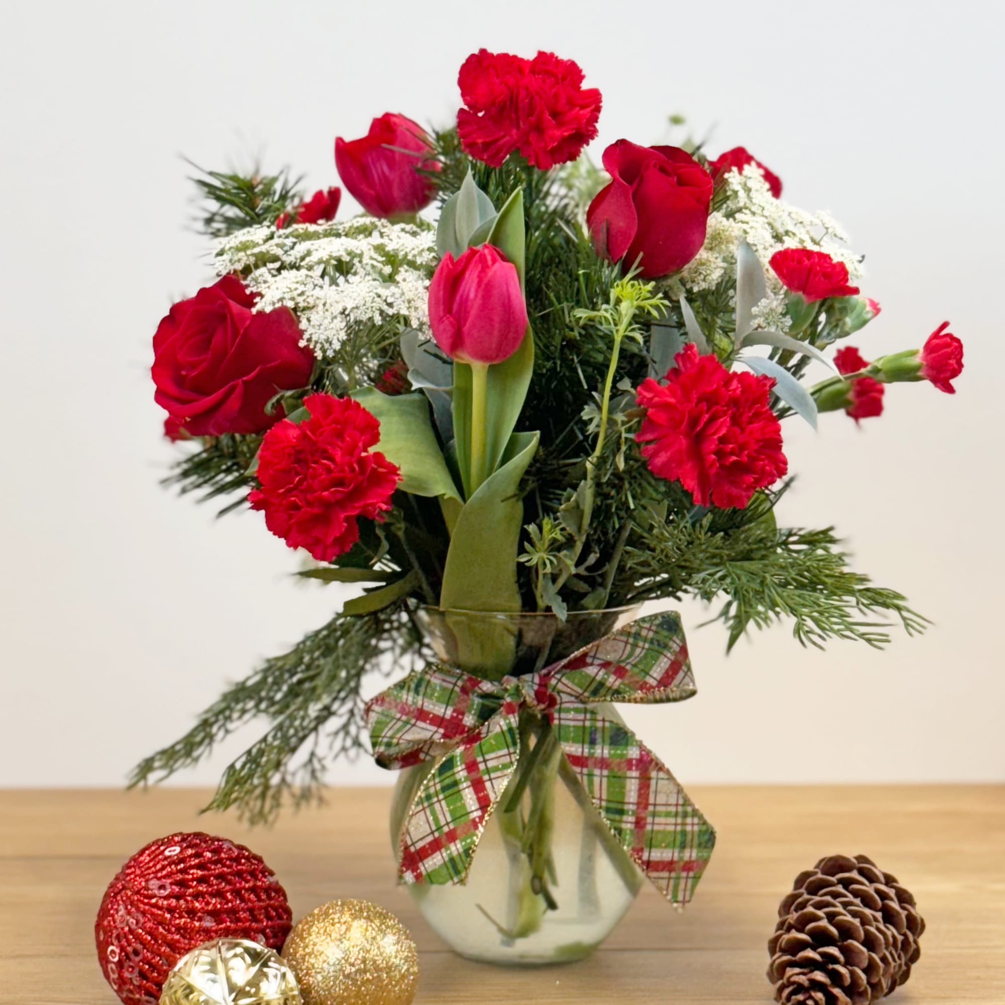Holiday bouquet of red roses, carnations, and tulips in a glass vase with plaid bow and ornaments.