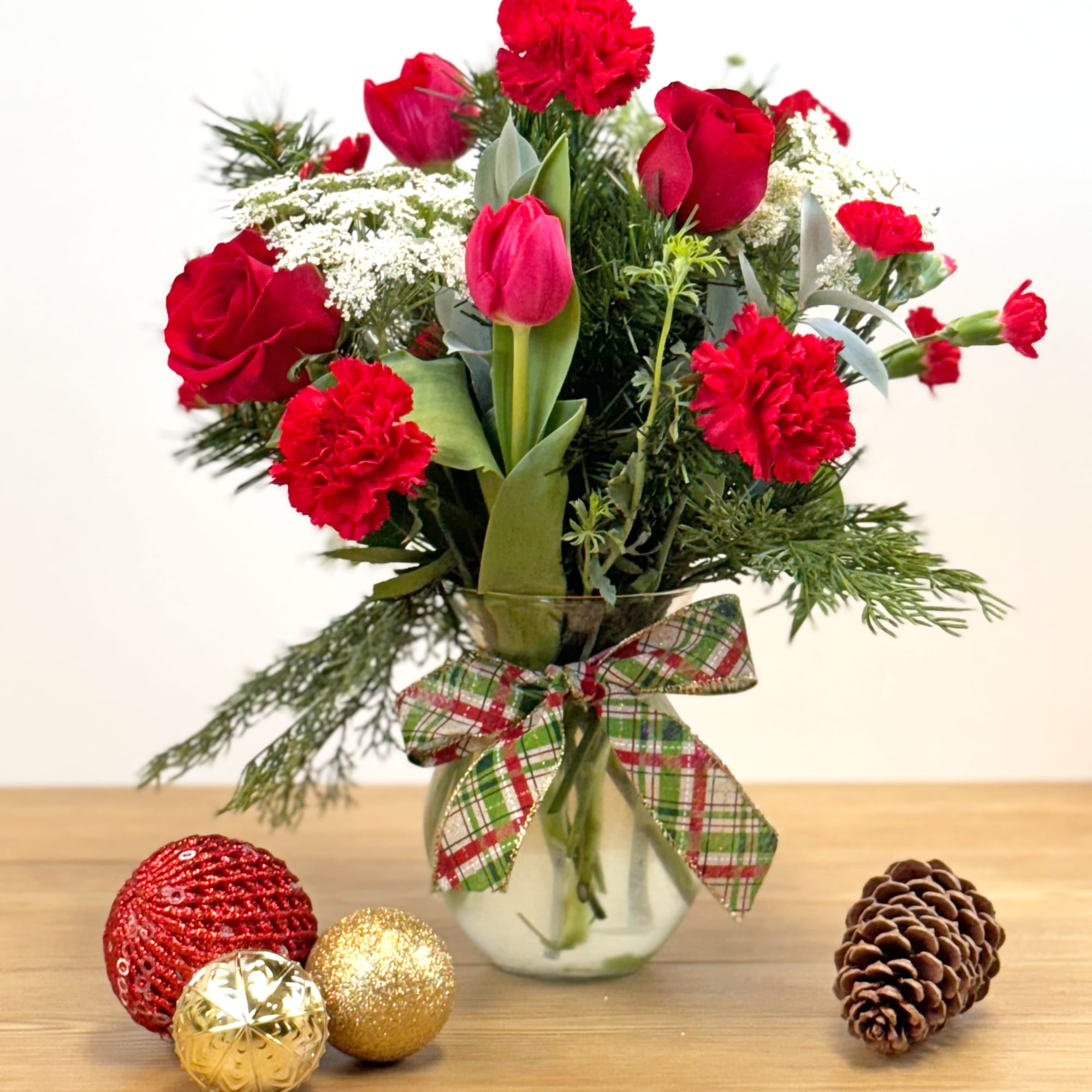 Red roses, carnations, and tulips in a glass vase with plaid bow, with ornaments and a pine cone on the table.