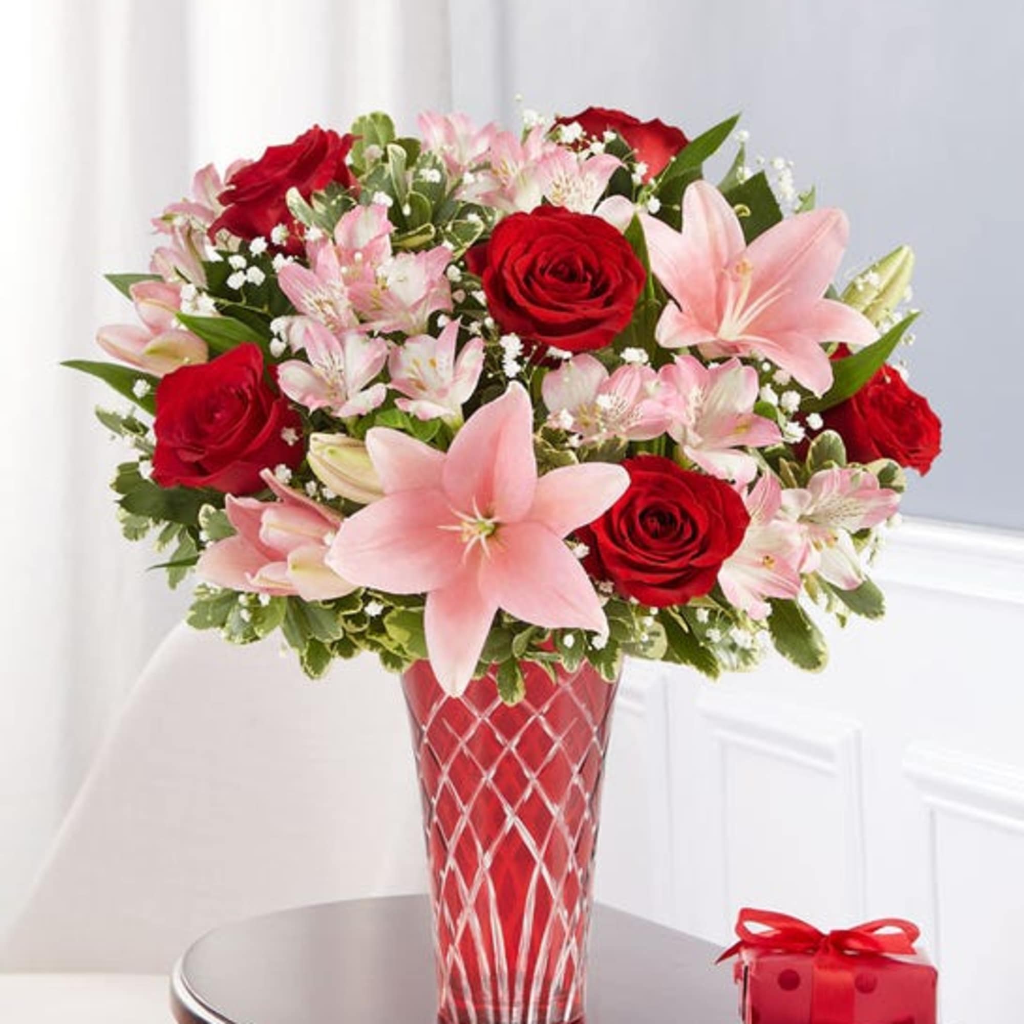 Red roses and pink lilies in a red glass vase with a gift box nearby