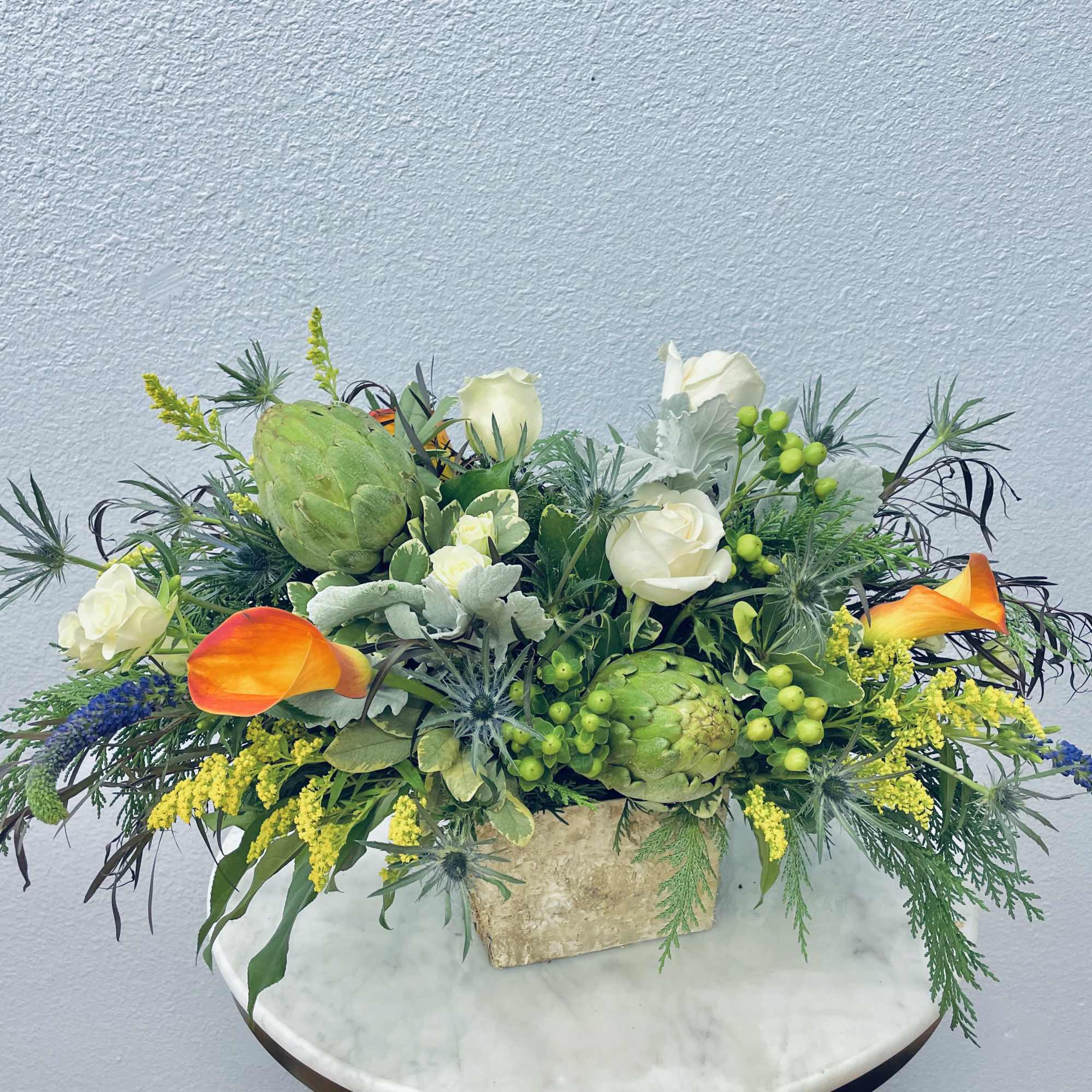 Mixed floral arrangement with white roses, orange calla lilies, and green artichokes in a stone vase