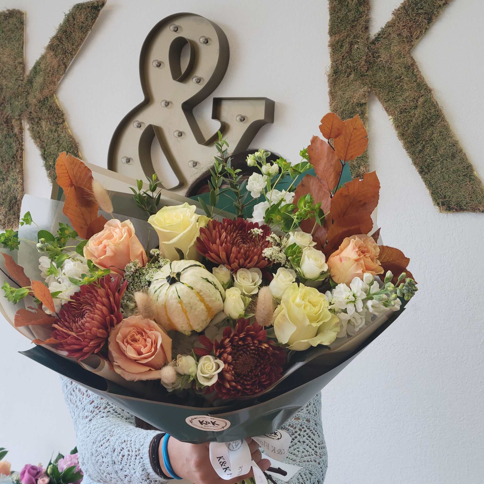 Handheld bouquet of roses, chrysanthemums, and a small pumpkin in dark wrap