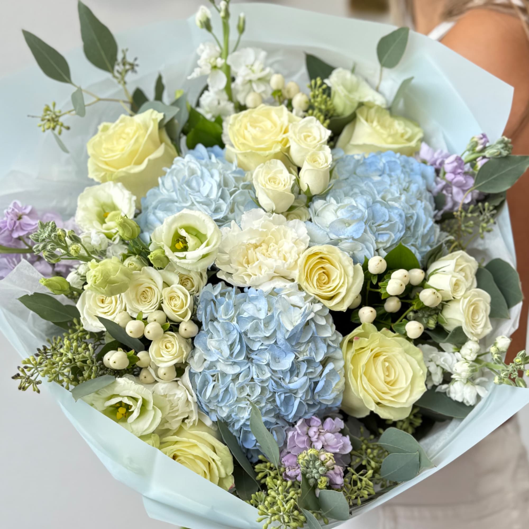 Bouquet of pale yellow roses, blue hydrangeas, and white flowers wrapped in light paper