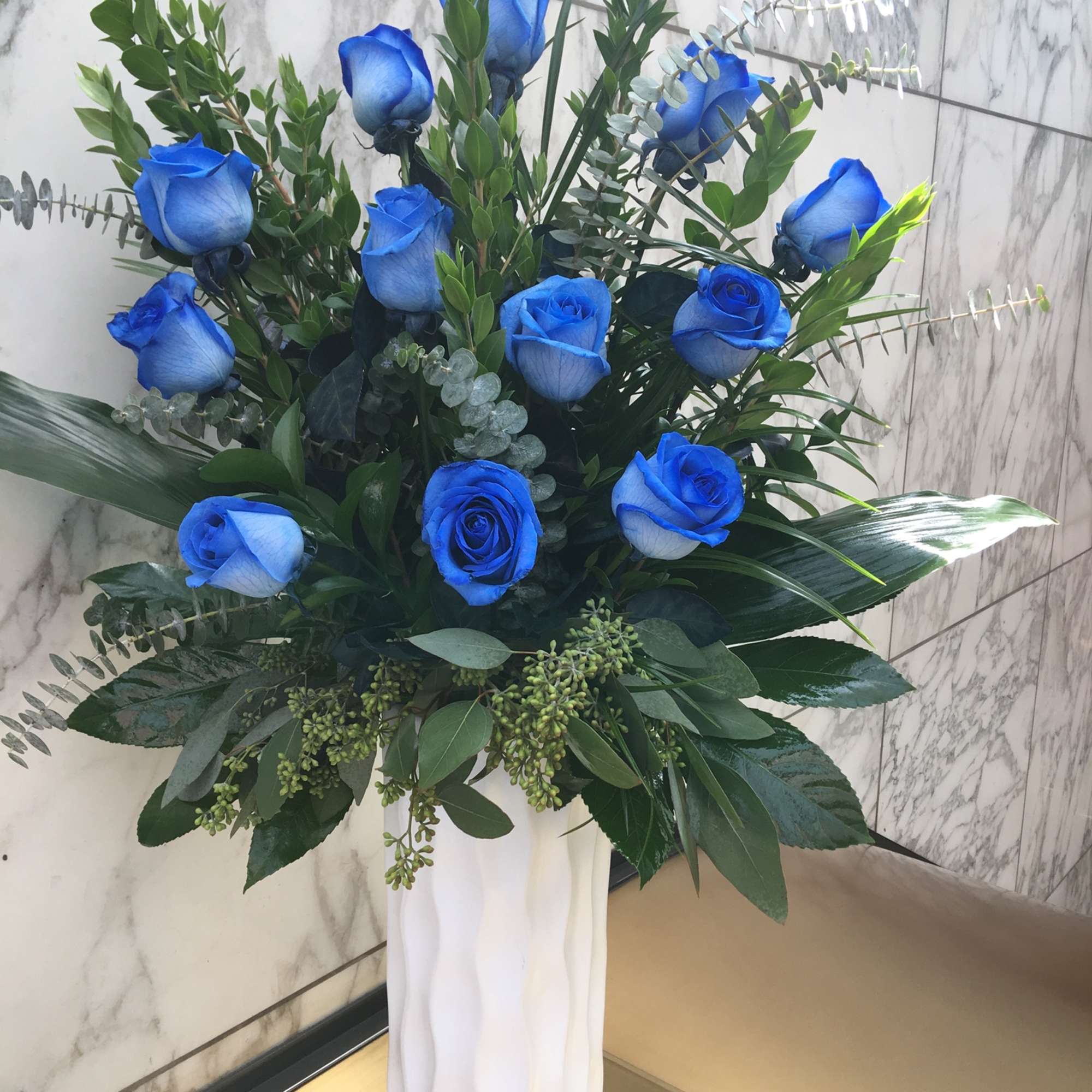 Blue roses in a tall white vase with mixed greenery