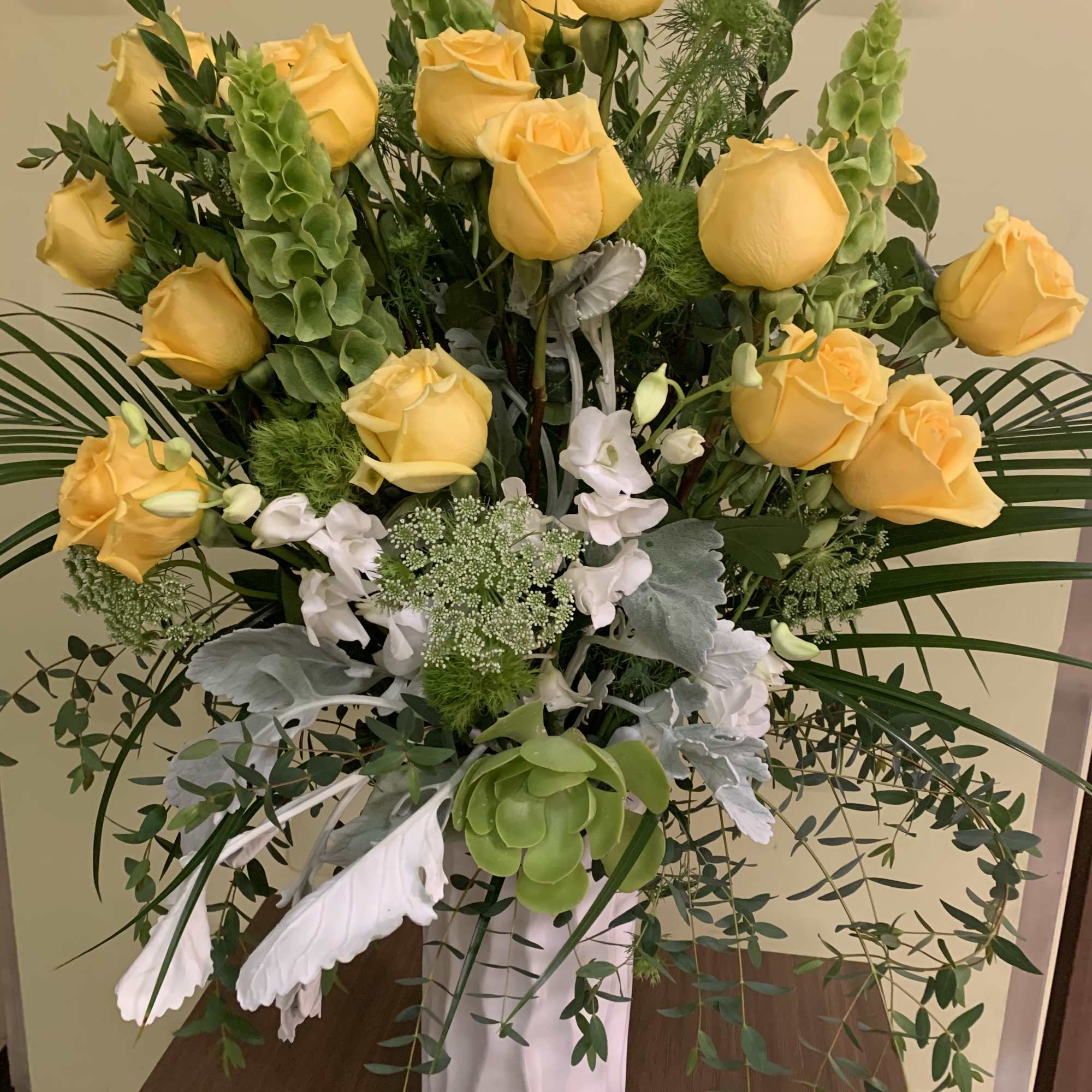 Yellow roses arranged in a tall white vase with white blooms and greenery