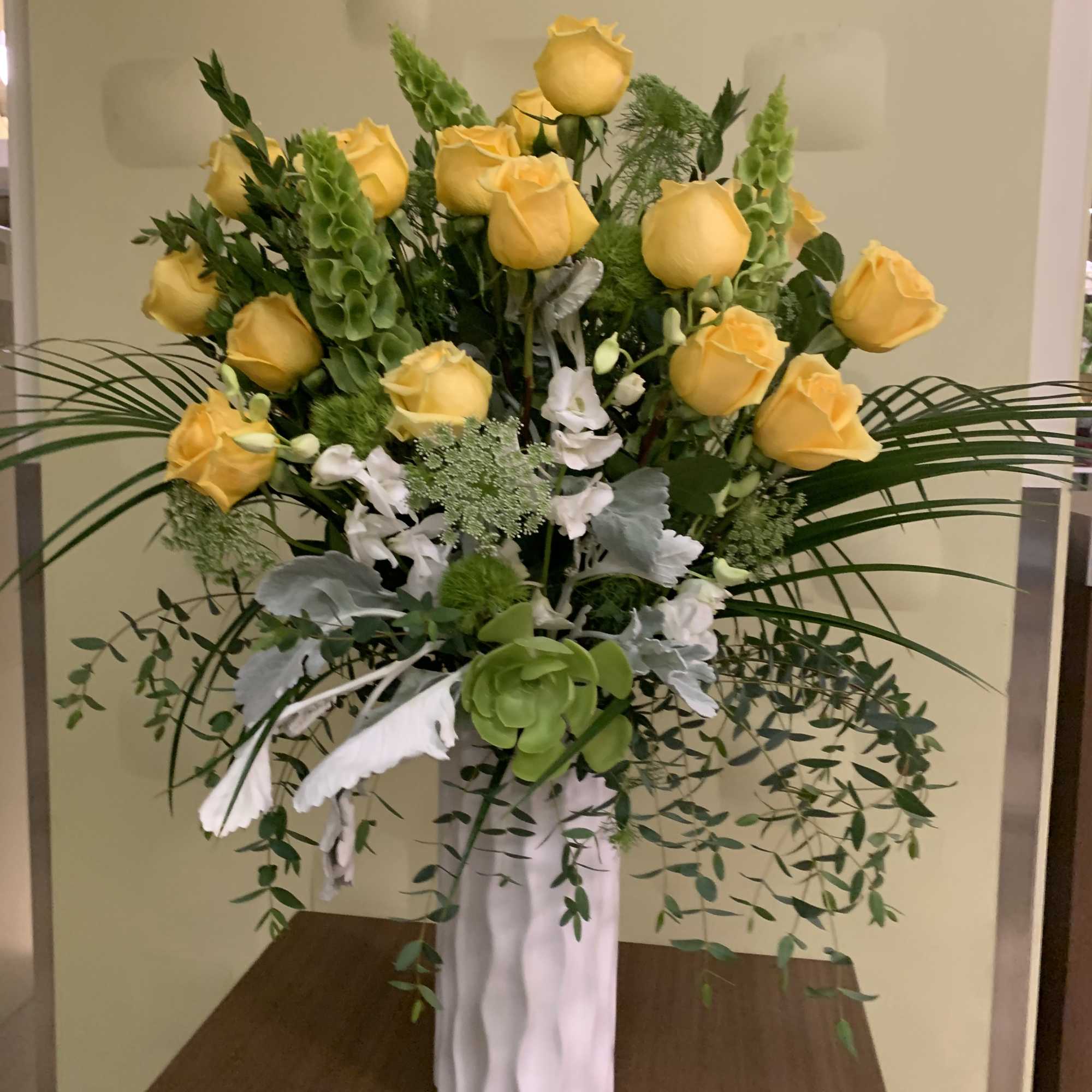 Yellow roses arranged in a tall white vase with greenery and white accents