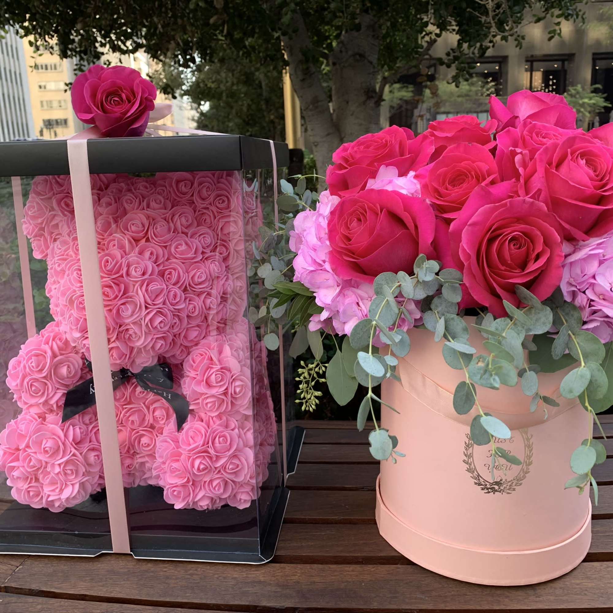 Pink rose teddy bear in a clear box beside a pink rose arrangement in a round hatbox