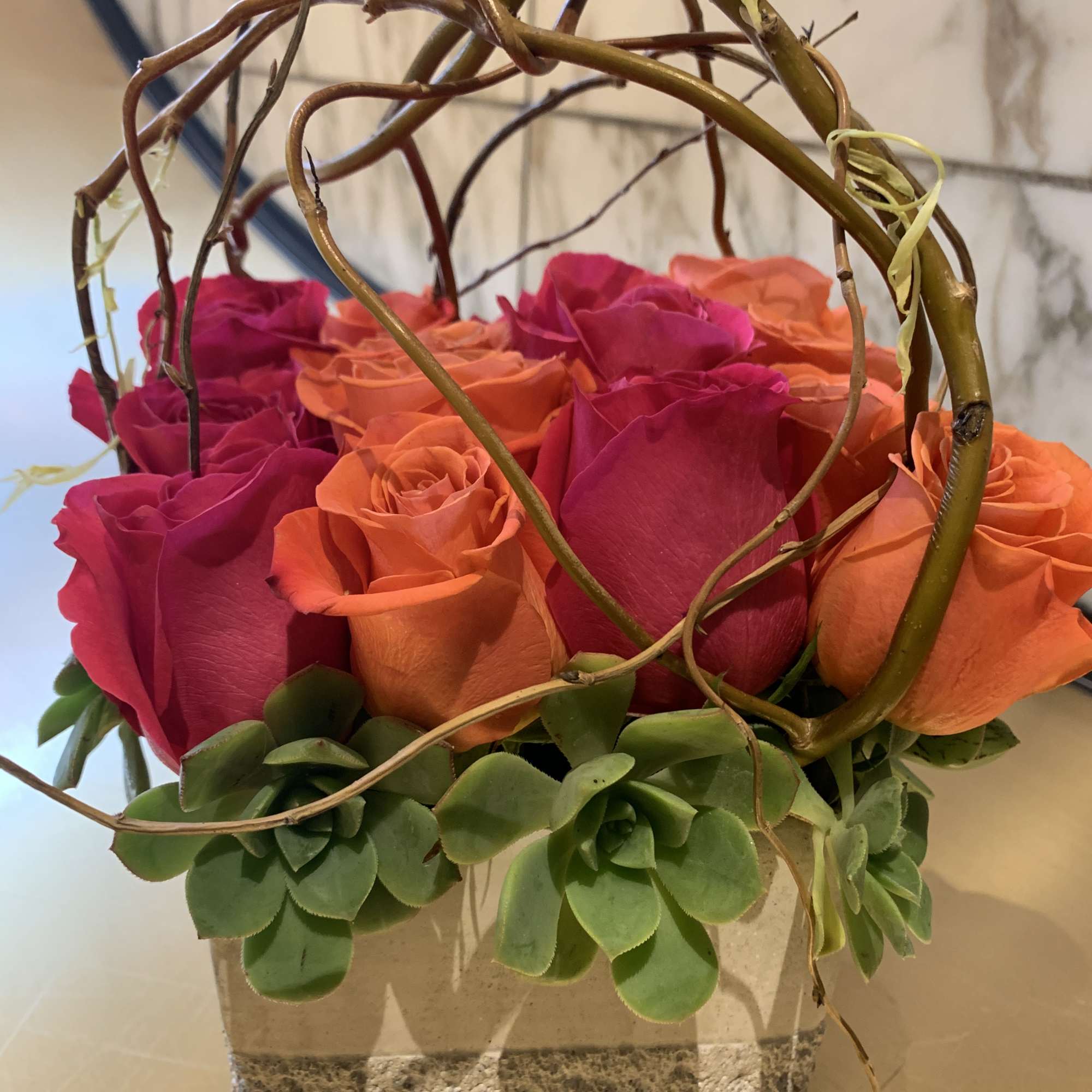 Pink and orange roses in a square stone planter with succulents and curly branches
