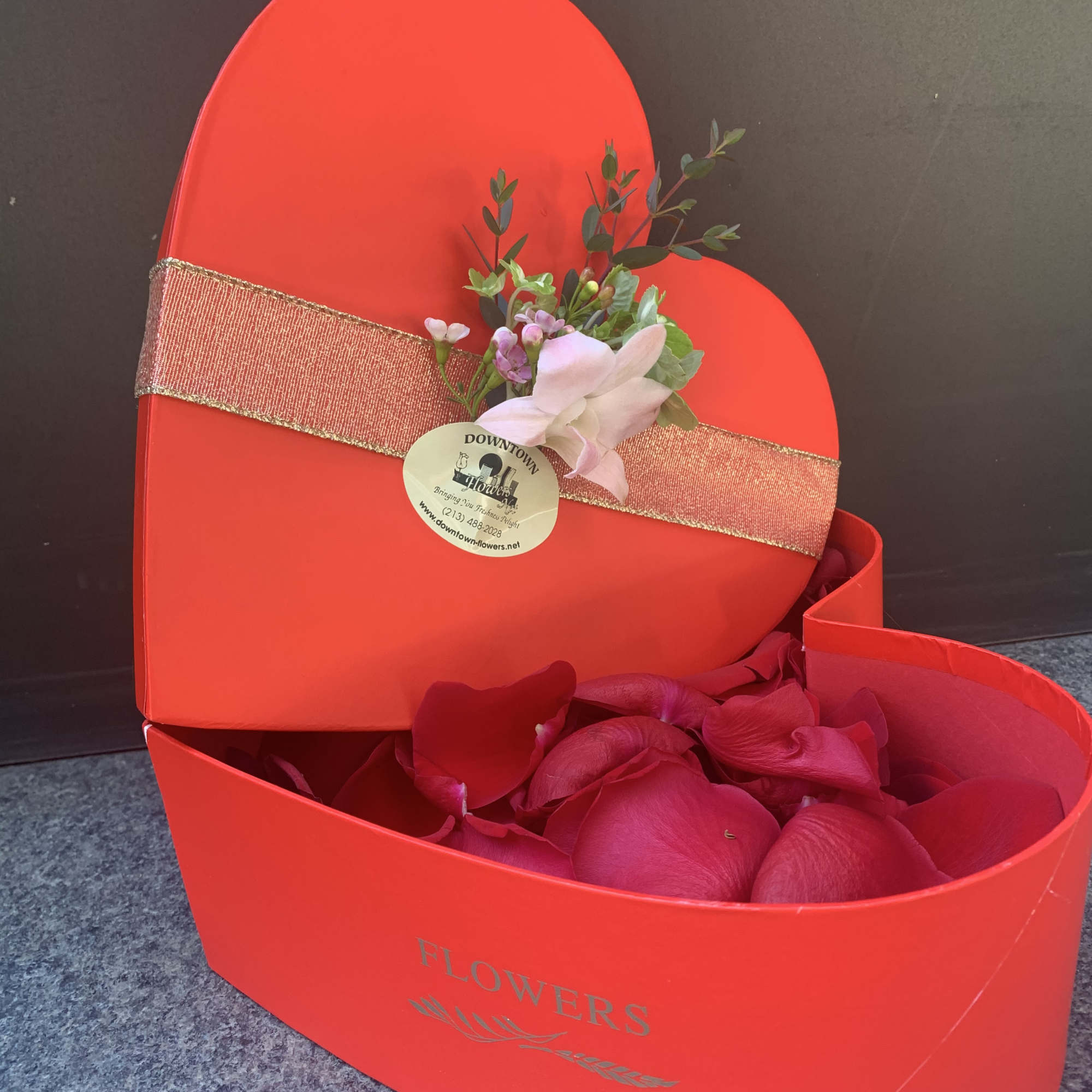 Red heart-shaped gift box with pink flower petals and a small floral accent