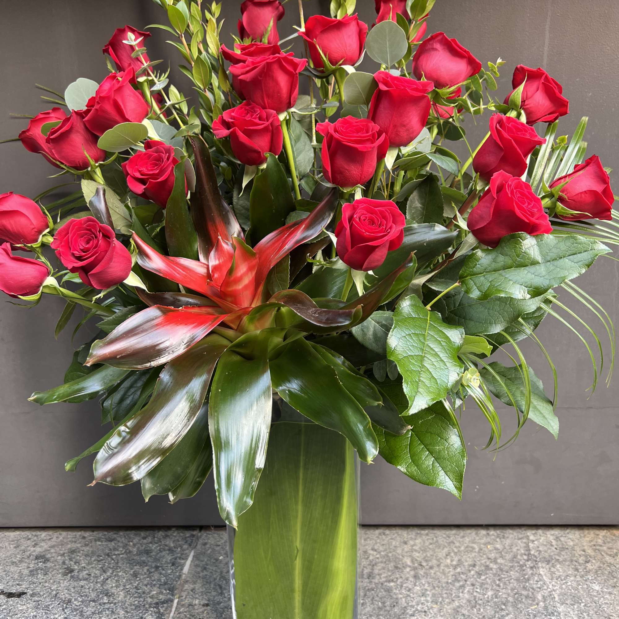 Red roses arranged in a tall glass vase with tropical foliage, photo 2 of 4