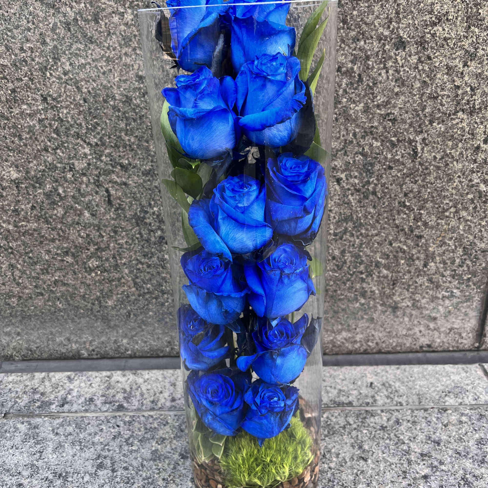 Tall clear vase with bright blue roses and green accents