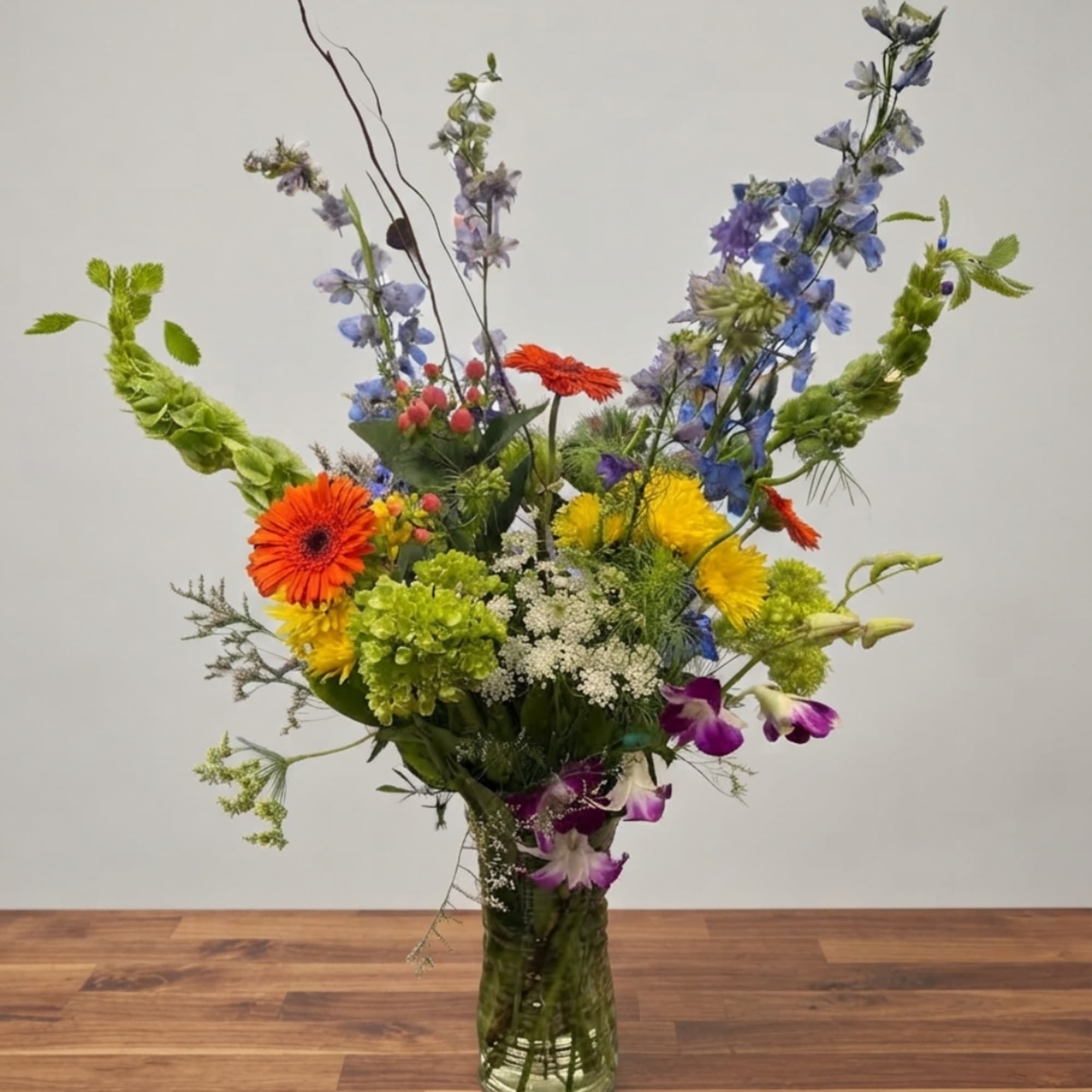 Tall mixed bouquet in a glass vase with orange, yellow, blue, and purple flowers.