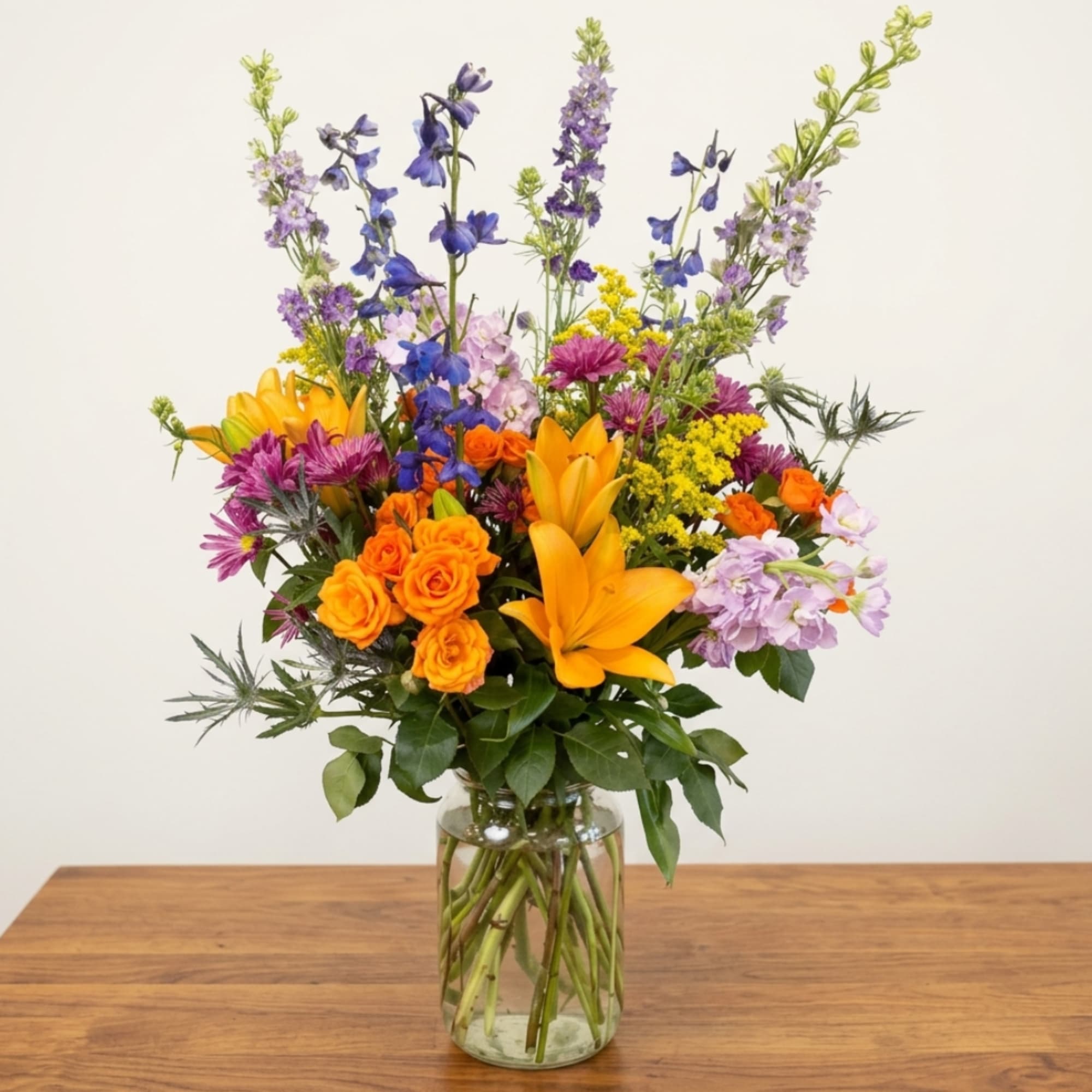 Mixed bouquet of orange roses, yellow lilies, and purple flowers in a glass vase