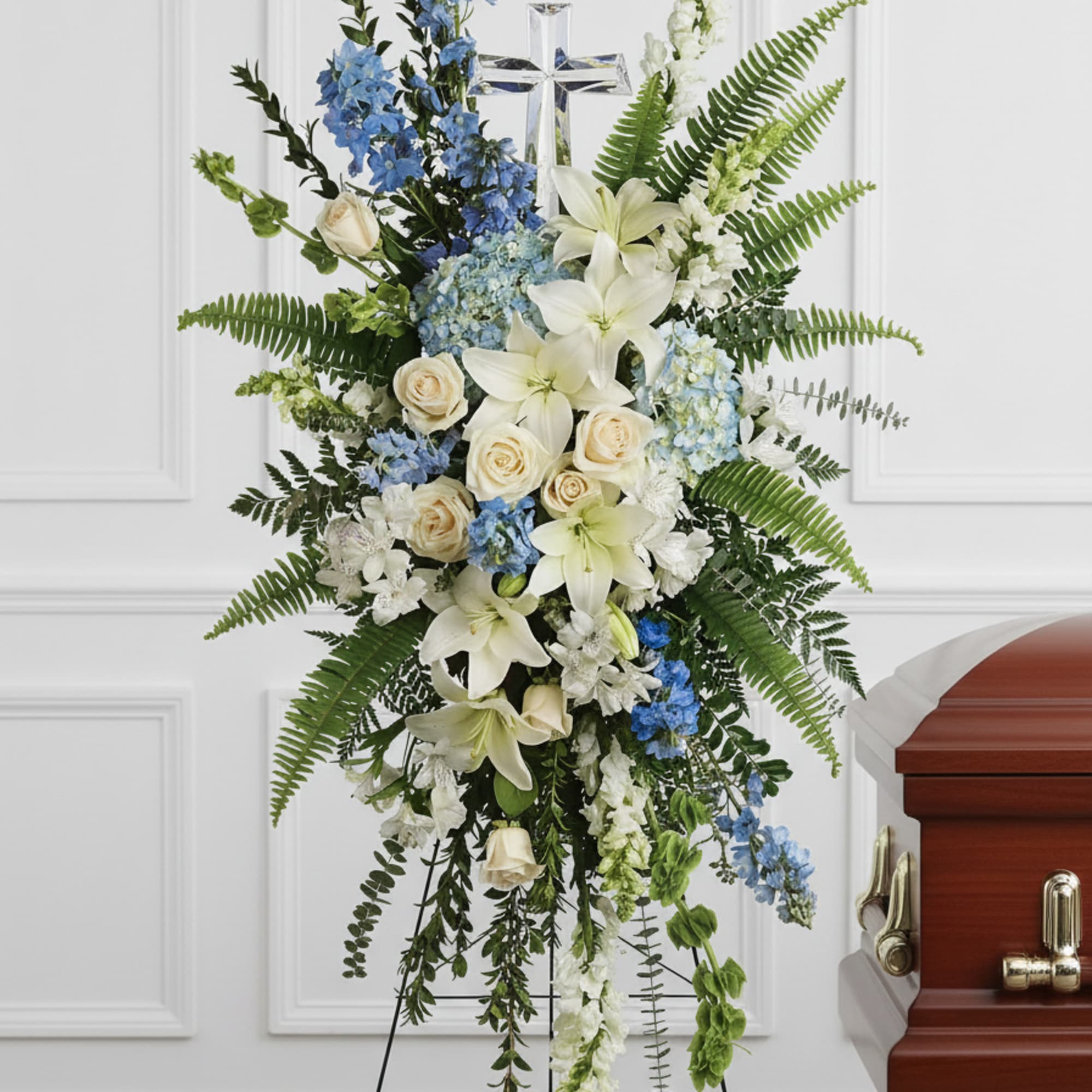 Tall standing spray of white lilies, cream roses, and blue flowers with a cross on an easel