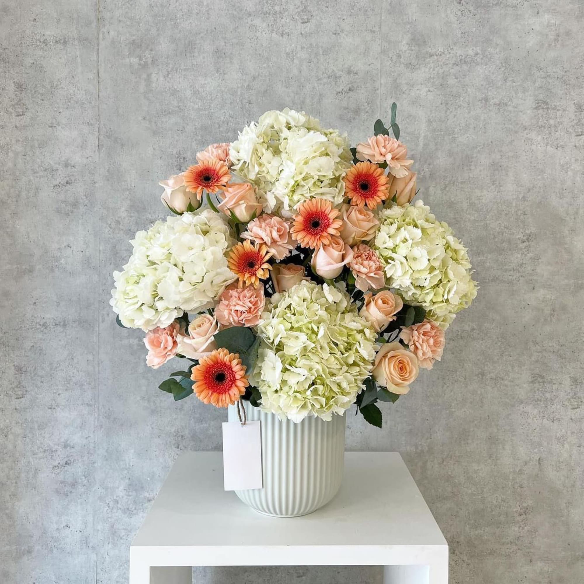 Peach roses and orange gerbera daisies in a white vase with hydrangeas