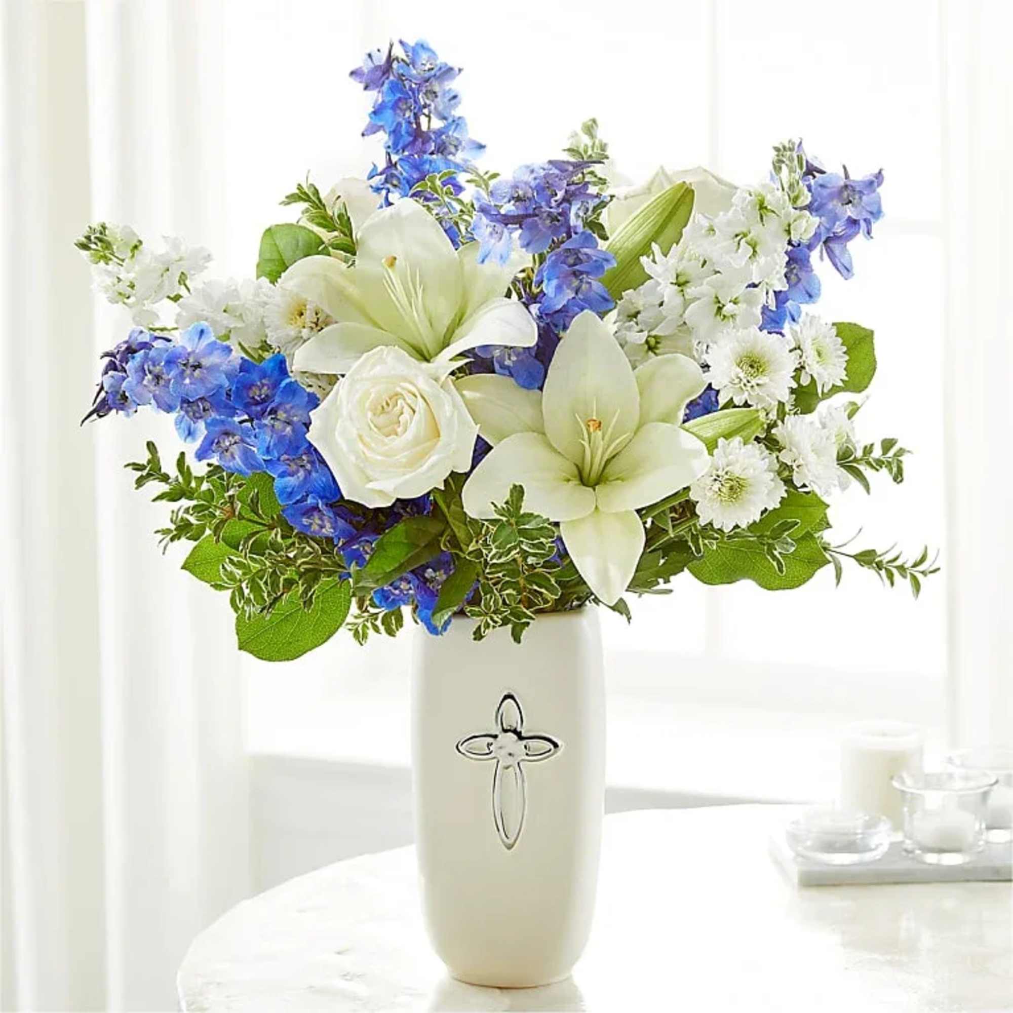 White lilies and blue delphinium in a white vase with a silver cross emblem.