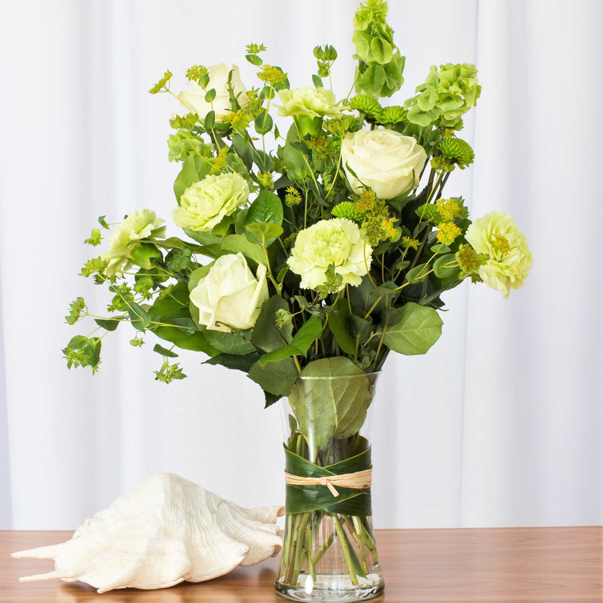Tall arrangement of pale green roses and carnations in a clear vase wrapped with leaves beside a seashell.
