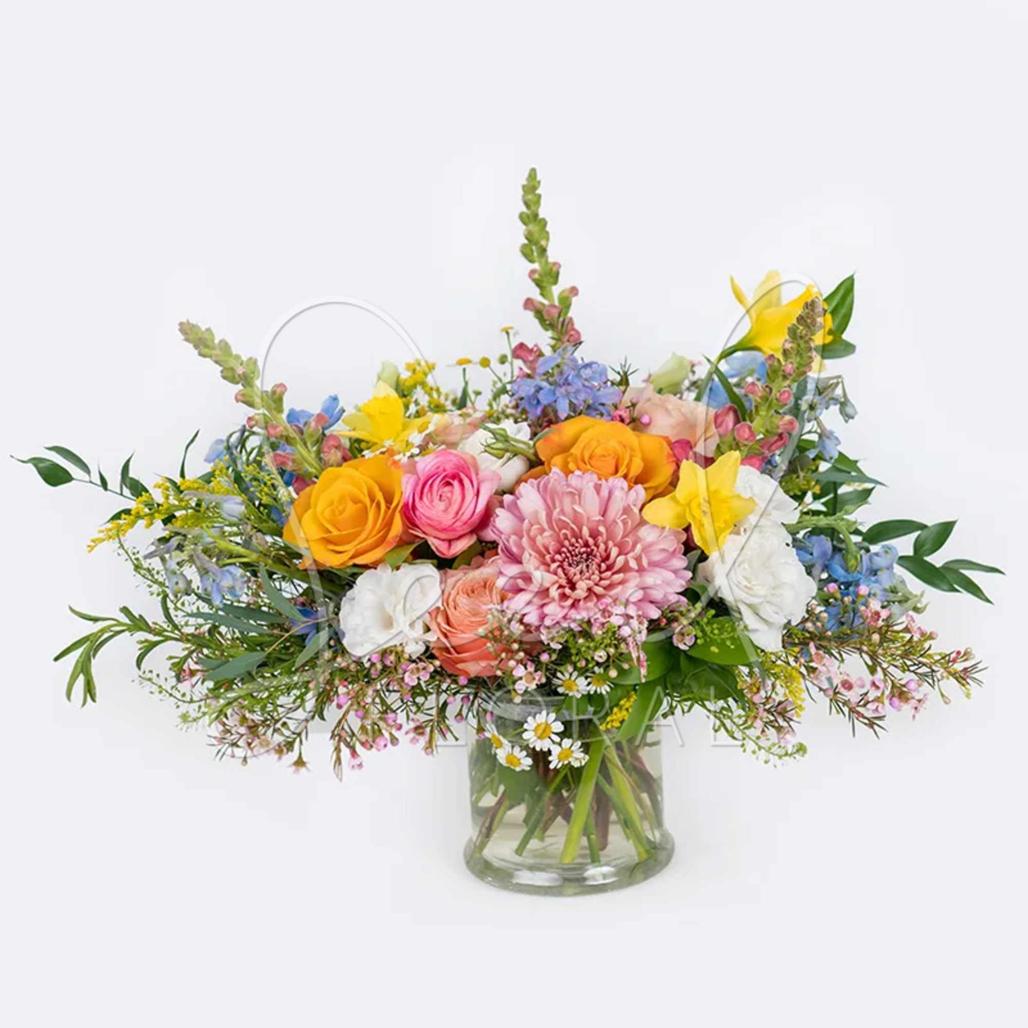 Low glass vase of mixed pink, yellow, white, and blue flowers in a lush garden-style arrangement