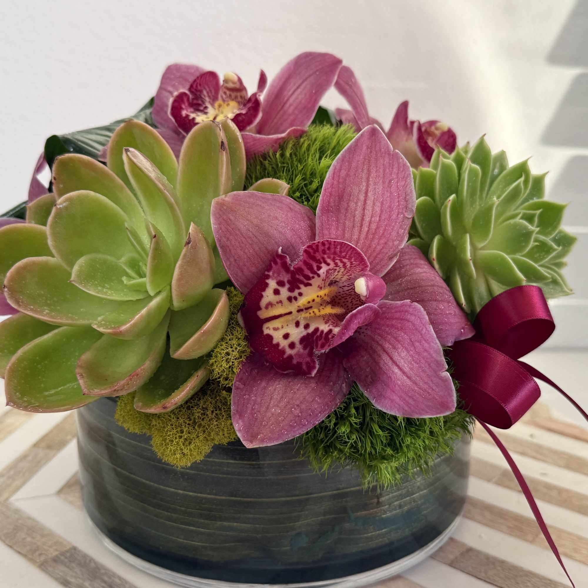 Pink orchids and green succulents arranged in a round black container with a ribbon