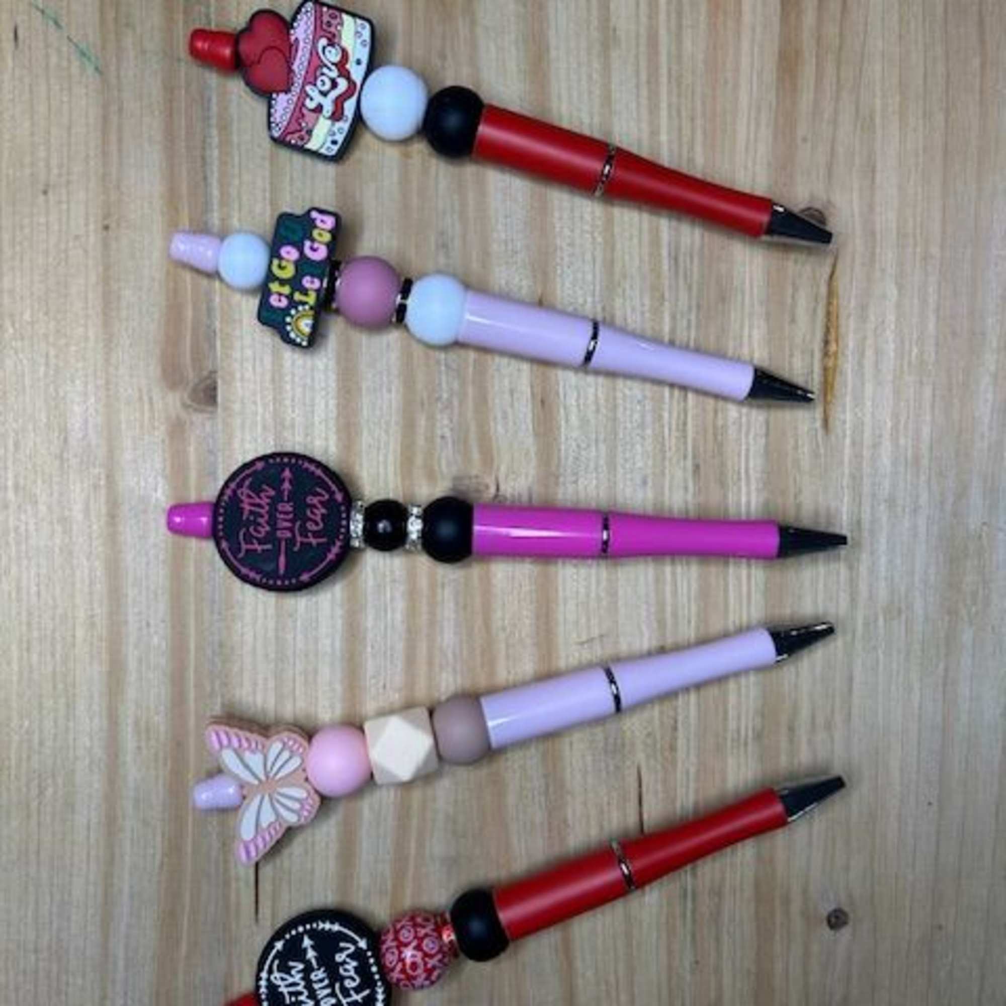 Five decorated pens with beads and charms arranged on a wooden surface.