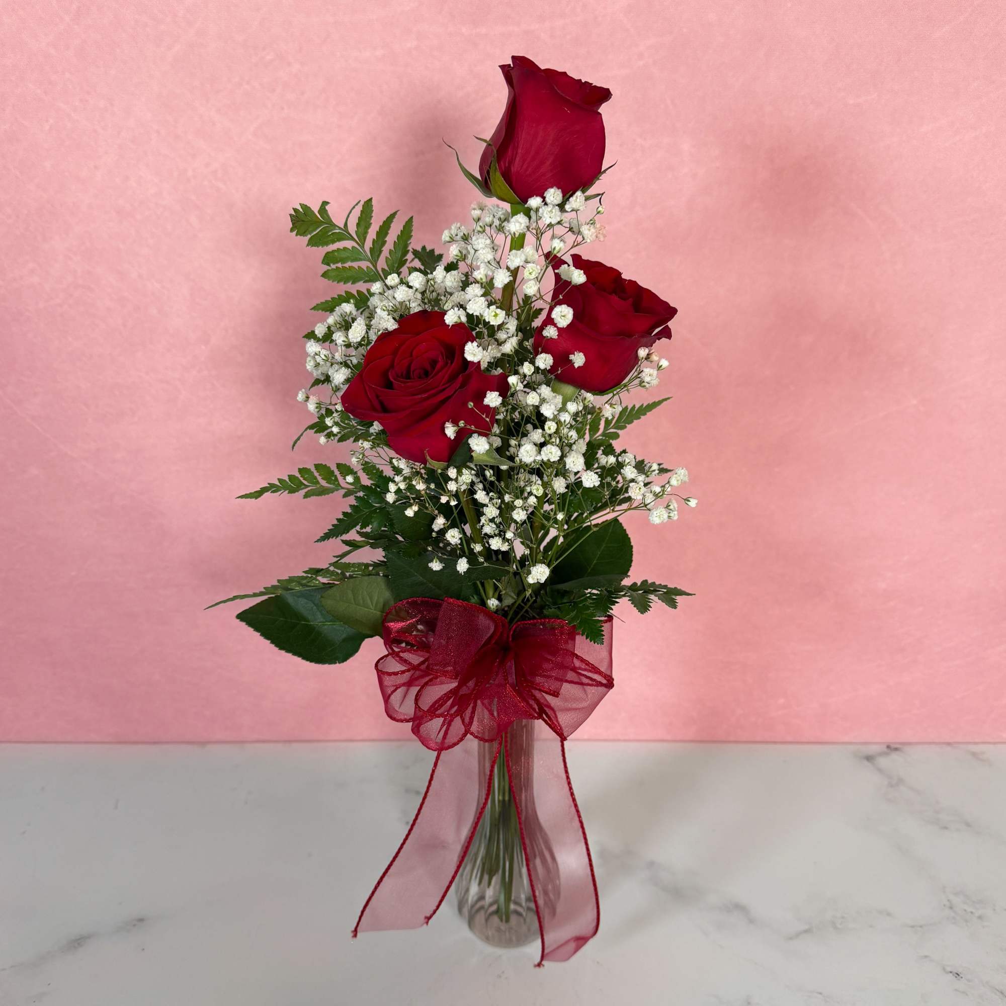 Three red roses in a clear vase with baby's breath and a red ribbon