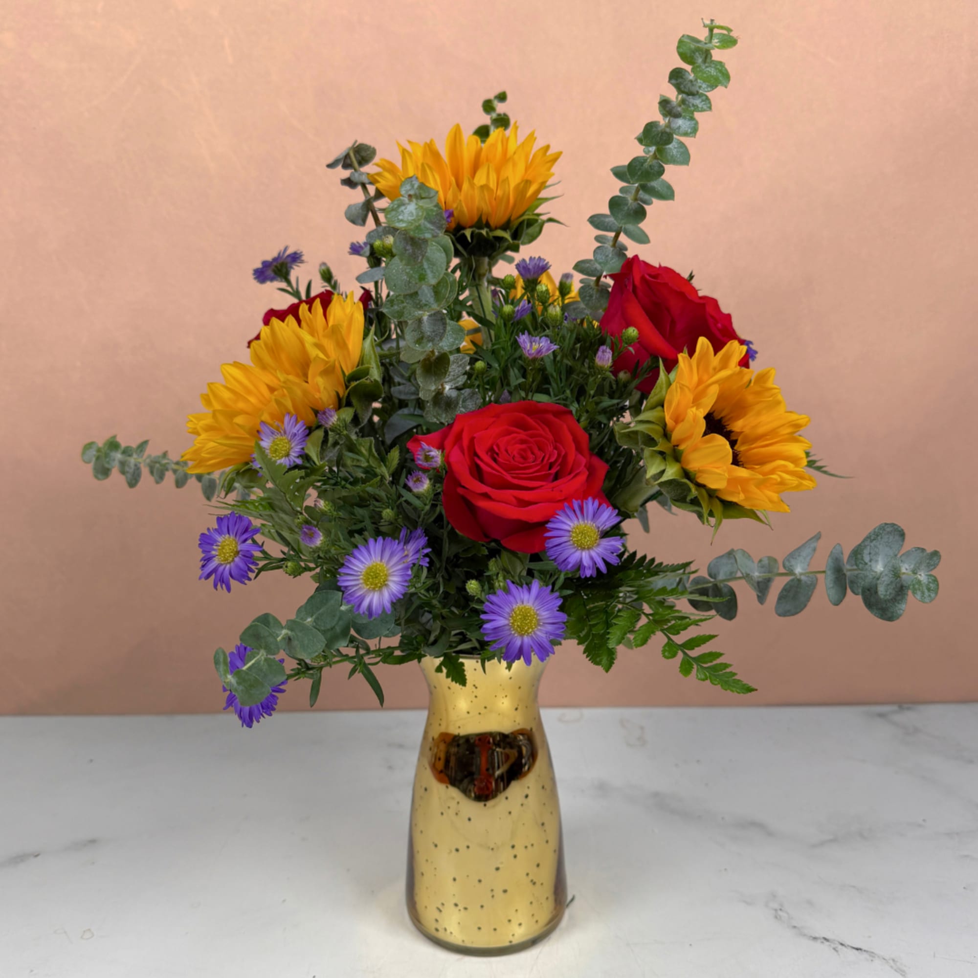 Mixed bouquet with red roses, yellow sunflowers, and purple daisies in a gold vase