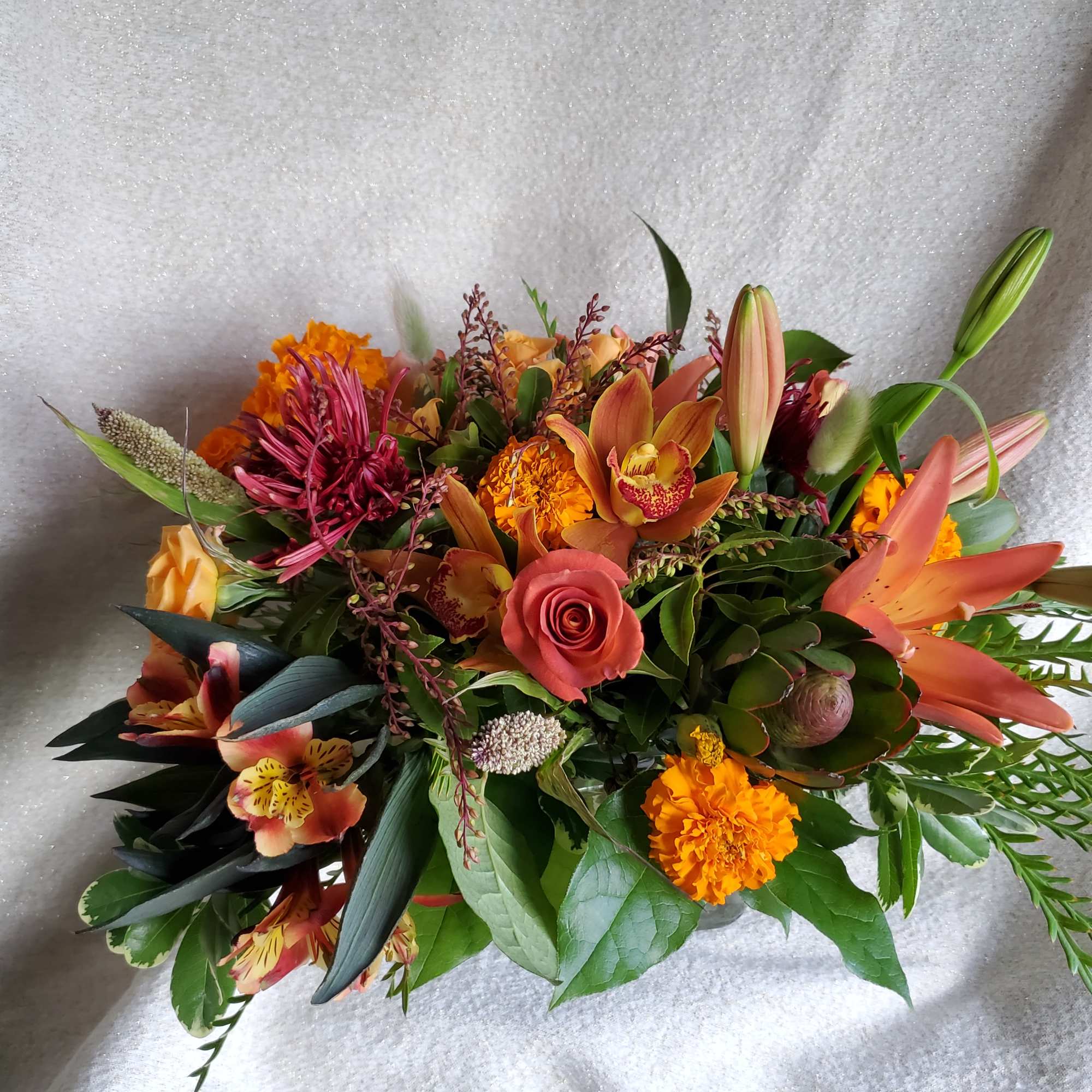 Low arrangement of orange lilies, roses, marigolds, orchids and mixed blooms in warm autumn tones