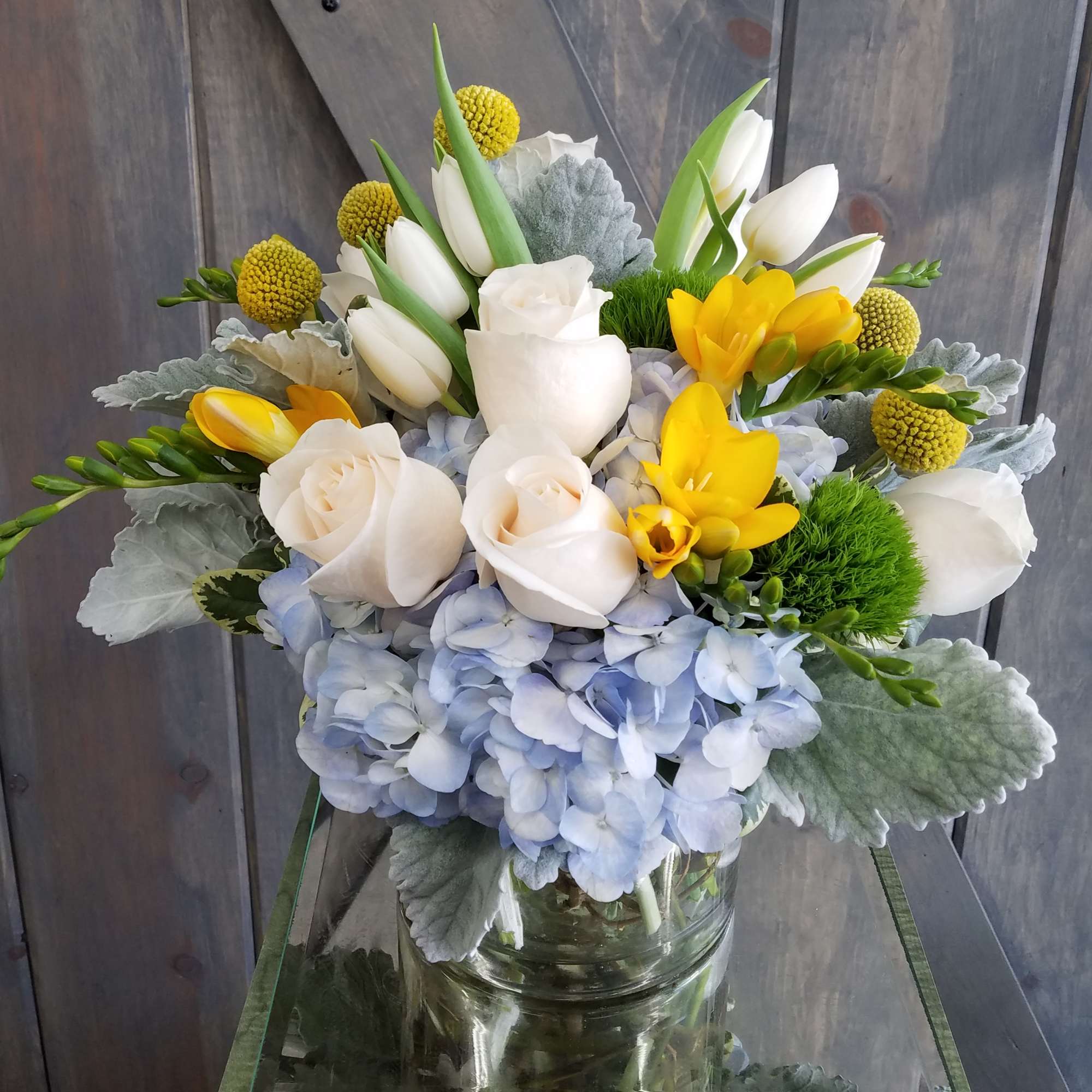Compact arrangement of white roses, tulips, yellow freesias, and blue hydrangeas in a clear glass vase