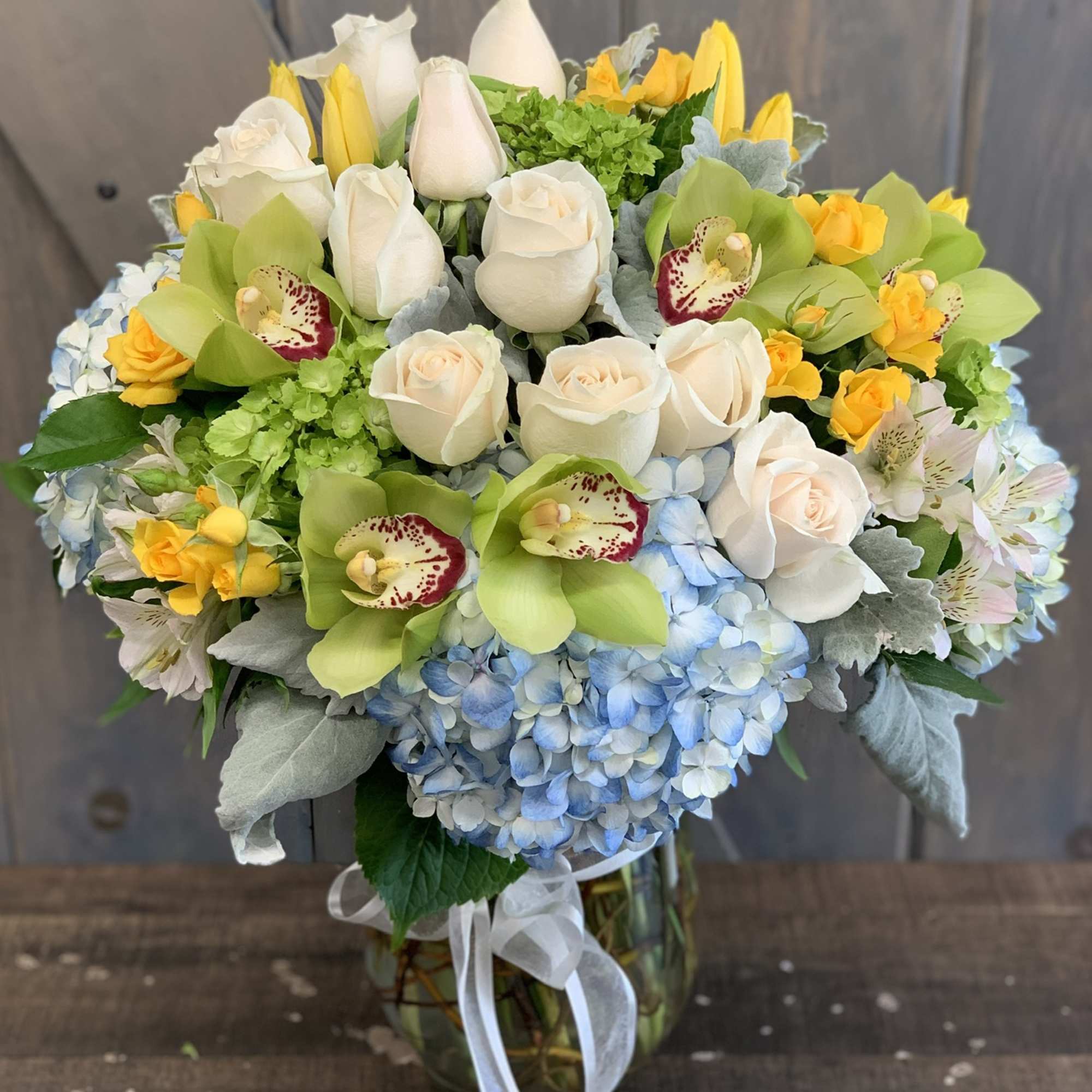 Premium arrangement of white roses, green orchids and yellow blooms over blue hydrangeas in a glass vase