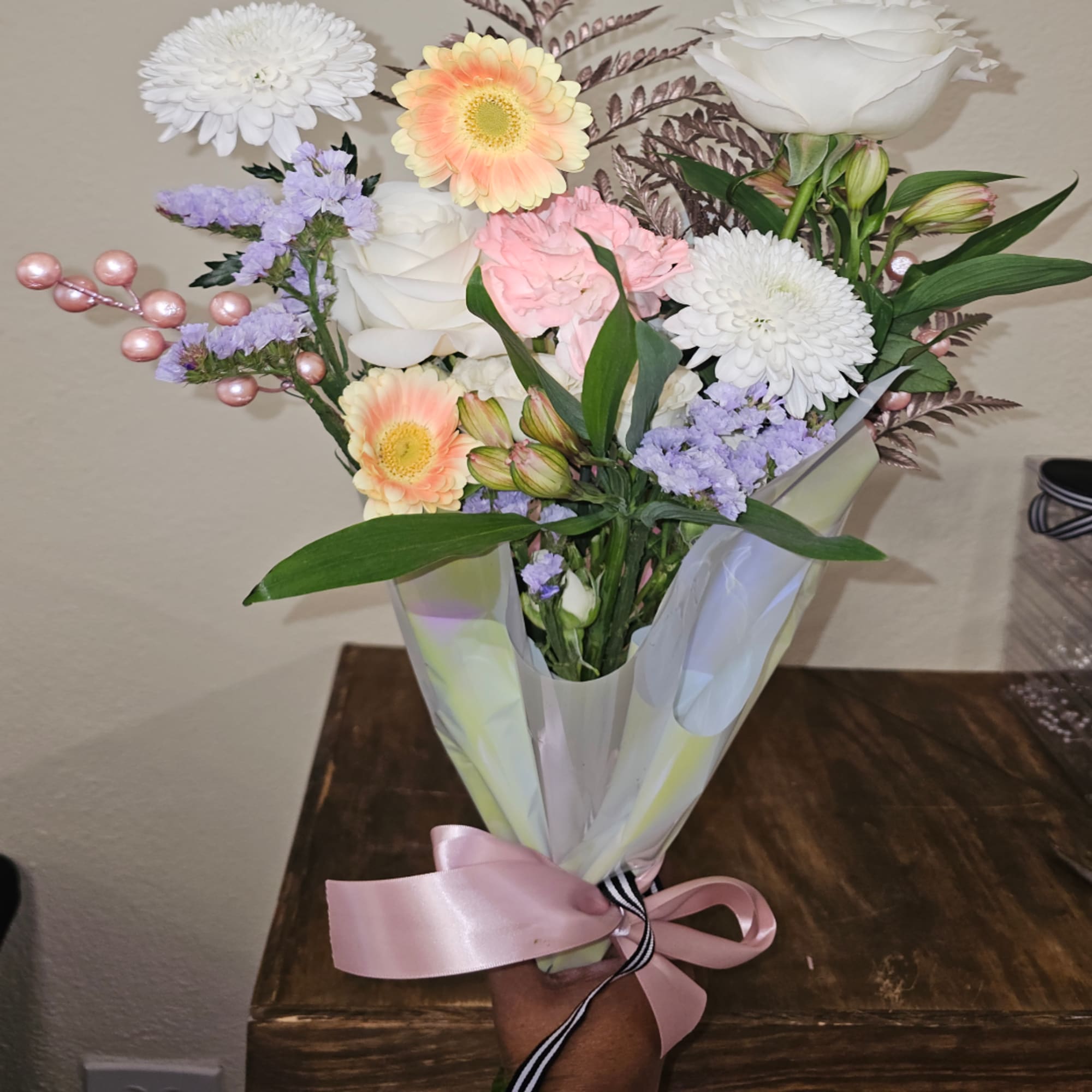 Handheld bouquet of white, pink, and peach flowers wrapped in paper with a pink ribbon