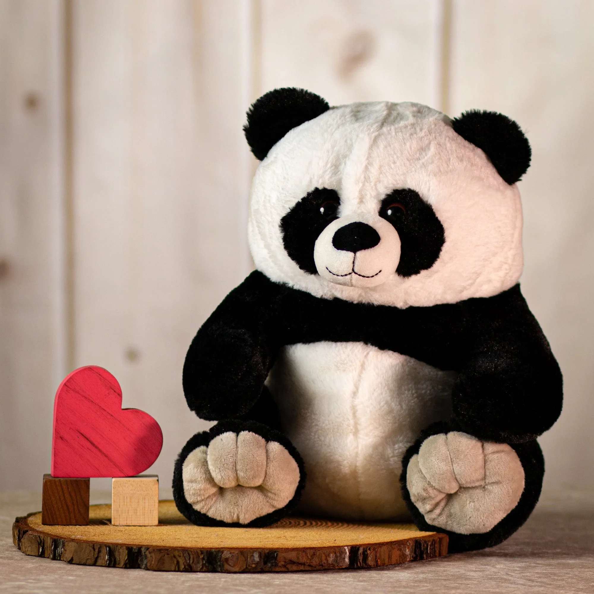 Plush panda bear sitting on a wood slab beside a small red heart ornament.