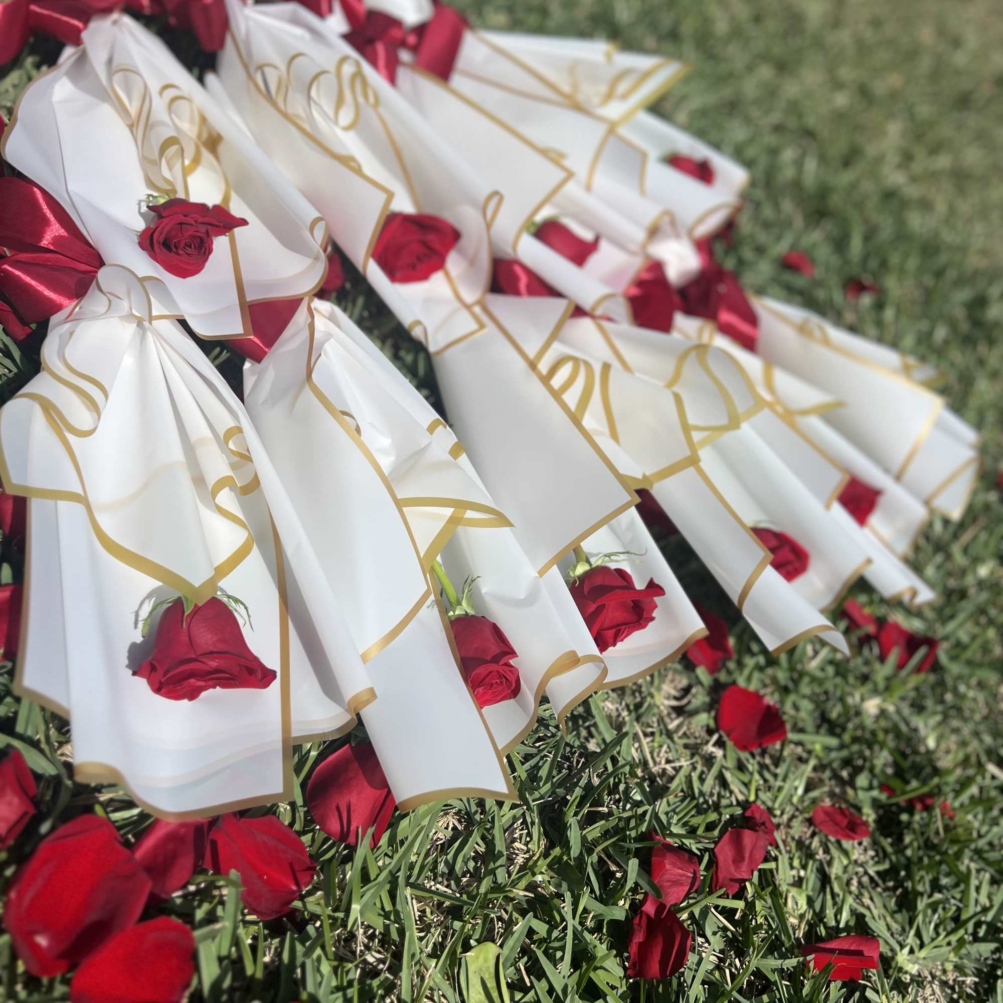 Red roses wrapped in white paper with gold trim and red ribbons on grass
