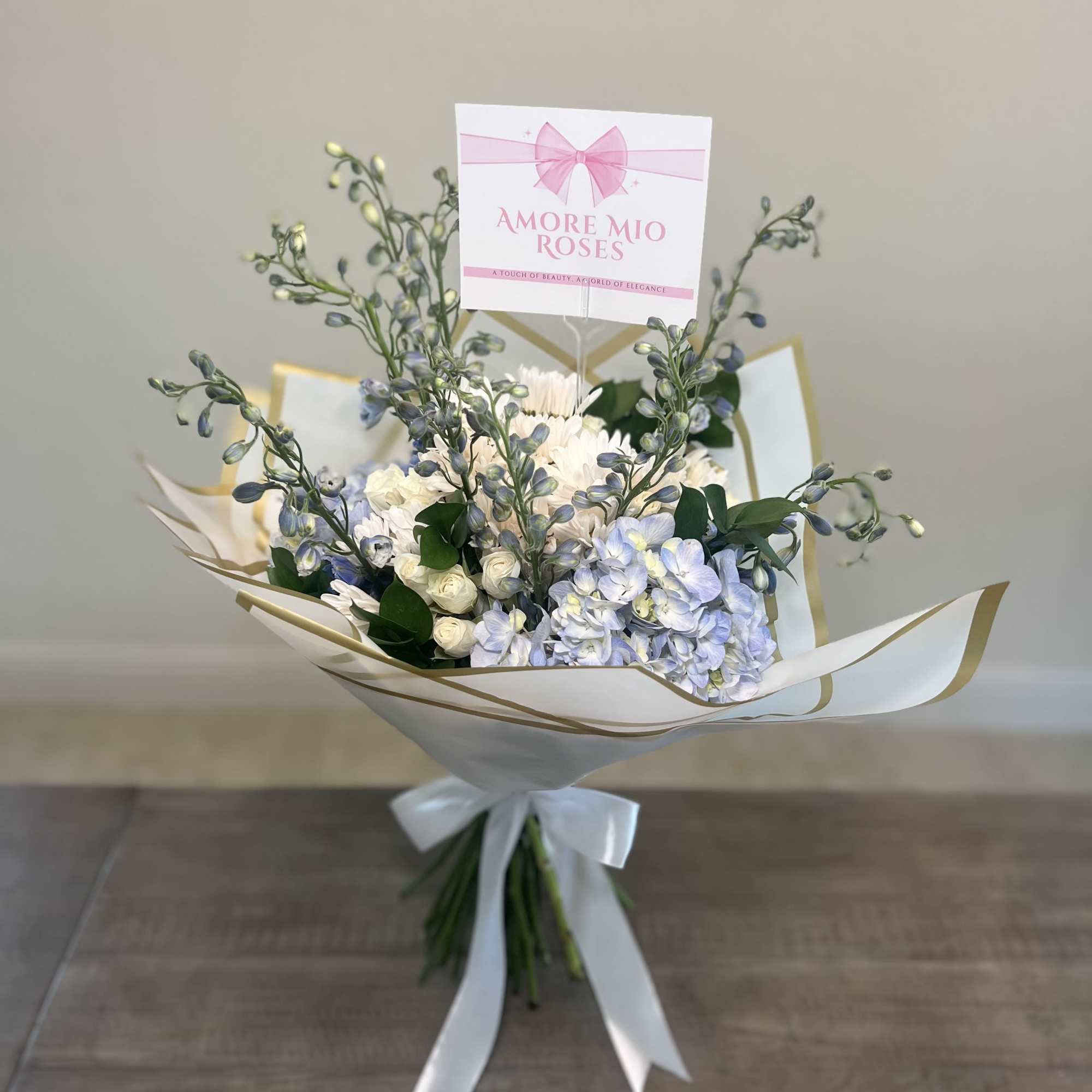 Bouquet of pale blue and white flowers wrapped in white paper with a pink card