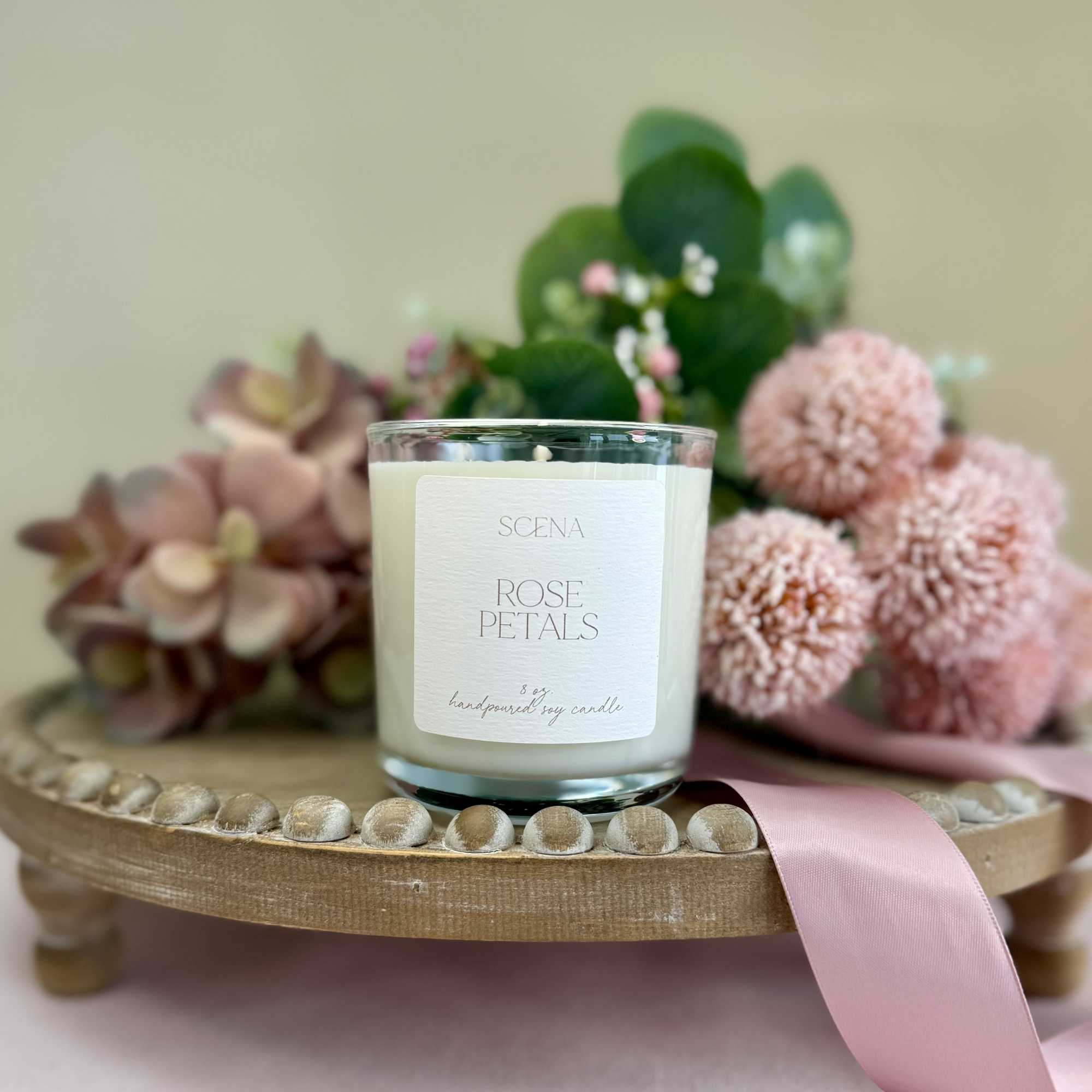 Rose-scented white candle in clear glass on wooden stand with pink flowers and ribbon